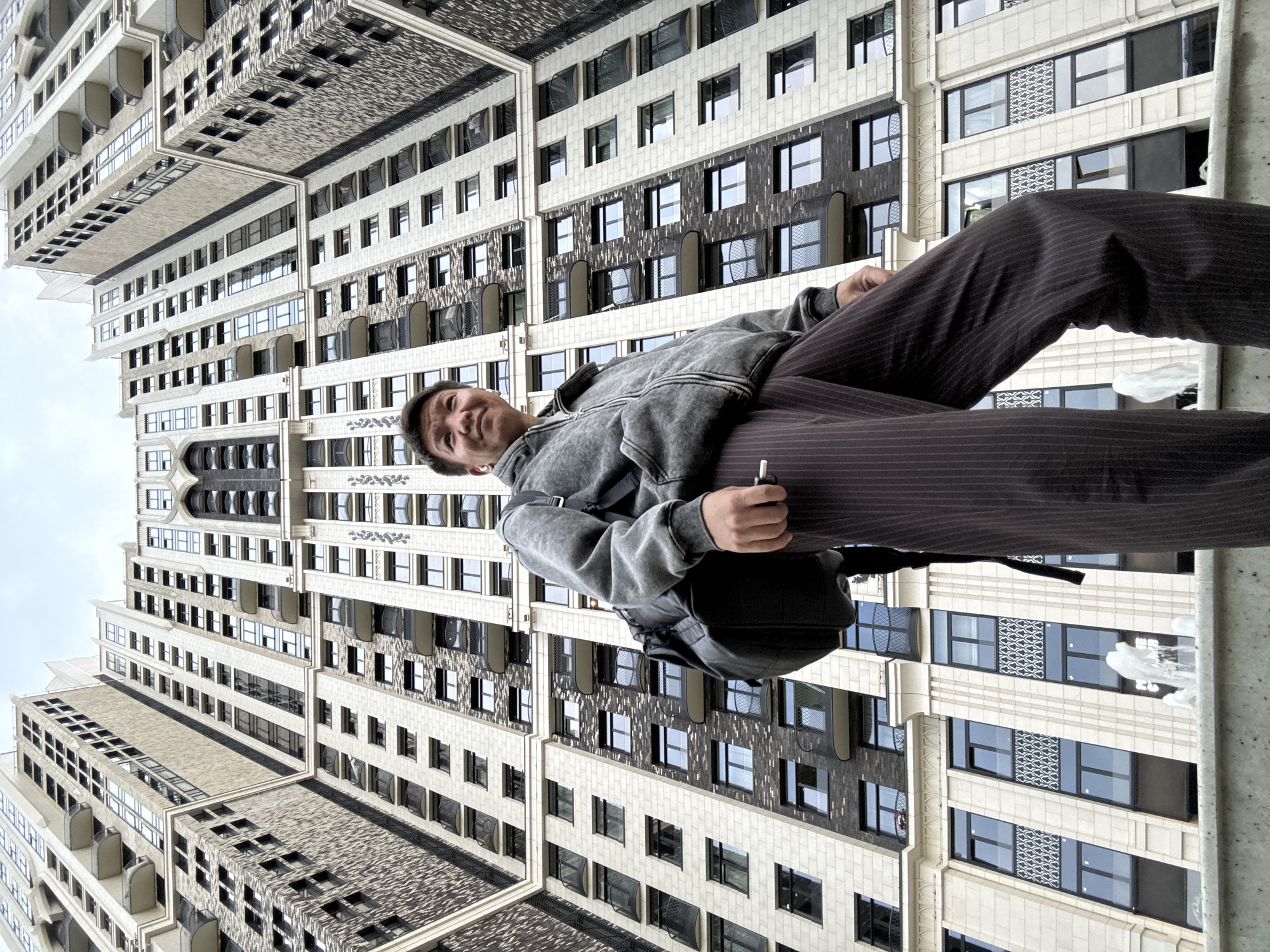 A low-angle medium shot captures a man standing in front of a large, modern building in Khimki, Russia. The building is tall and features many windows and balconies, with a pale beige and dark grey facade. The man, viewed from the waist up, is wearing a grey hooded jacket, dark pinstriped pants, and a black backpack. He is looking upwards and to his left, with a cigarette in his right hand. A small fountain is visible in the foreground. The sky is overcast.