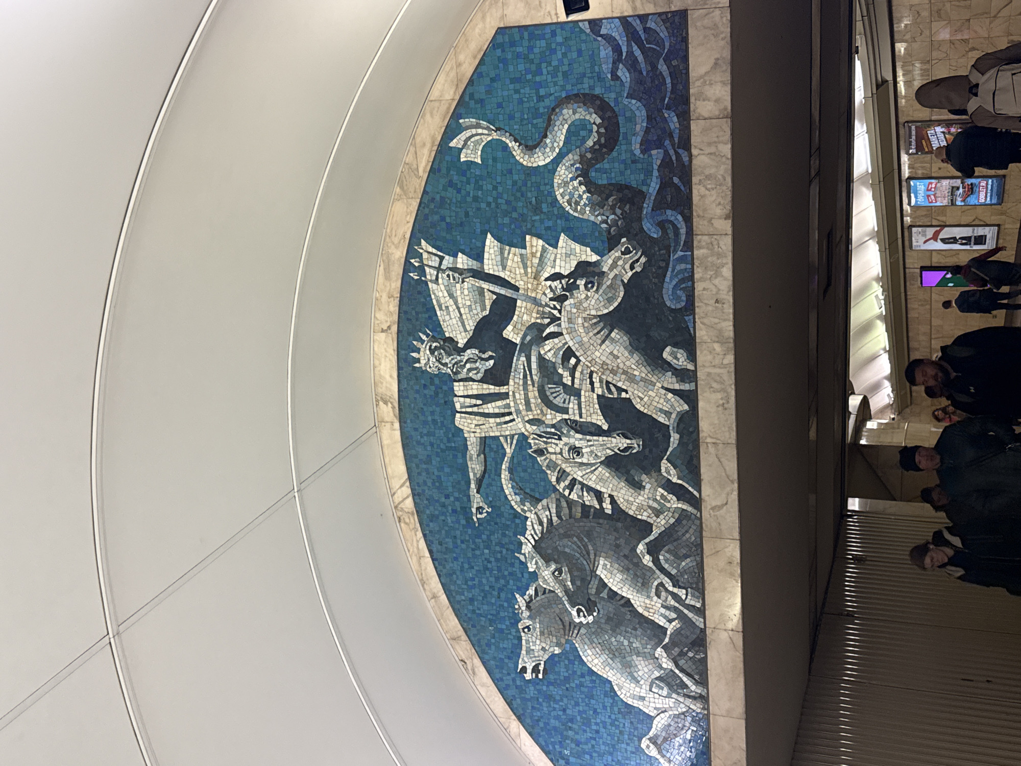 A mosaic depiction of Poseidon in the Saint Petersburg Metro. The mosaic depicts the god in a chariot pulled by horses, fighting a sea serpent. People walk nearby. Advertisements adorn the walls.