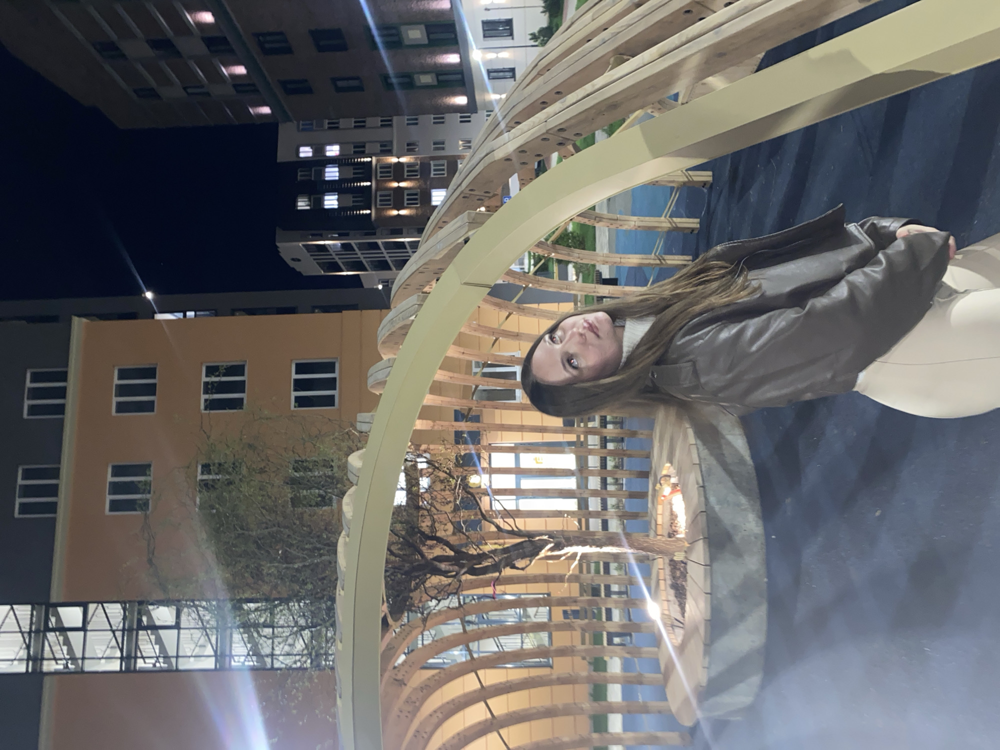 The image shows a woman standing within a large, arched wooden structure, likely an art installation or architectural feature, located in Pervomaysky, Russia. The structure is composed of multiple curved wooden beams arranged in a semi-circular form, giving the impression of a ribcage or skeleton-like construction. In the background, various buildings are visible, including high-rise residential apartments with numerous windows illuminated at night. One building, an orange color, has a pedestrian walkway spanning between its two structures. A tree with sparse branches is also visible. The woman is wearing a brown jacket and light-colored pants. The overall lighting suggests it is nighttime. A light source from the ground highlights the lower portion of the structure.