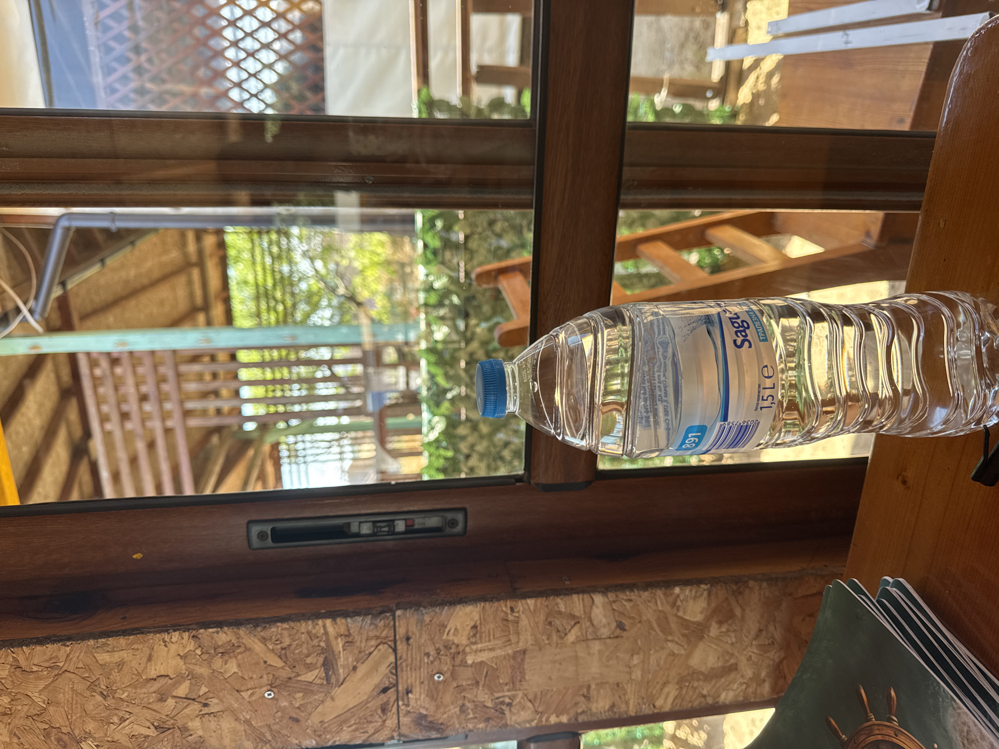 The image captures an interior view through a wooden-framed window, likely from a building in Sozopol, Bulgaria, based on the "Sagu" water bottle labeled "ТРАПЕЗНА В" ("Table Water") which is a Bulgarian brand. The window looks out onto a partially obscured exterior featuring greenery and architectural elements. A 1.5-liter "Sagu" water bottle with a blue cap is placed on a wooden surface near the window. Adjacent to the bottle are green items with an image of a ship's wheel. The interior construction consists of plywood. A sliding window lock is visible above the plywood section.