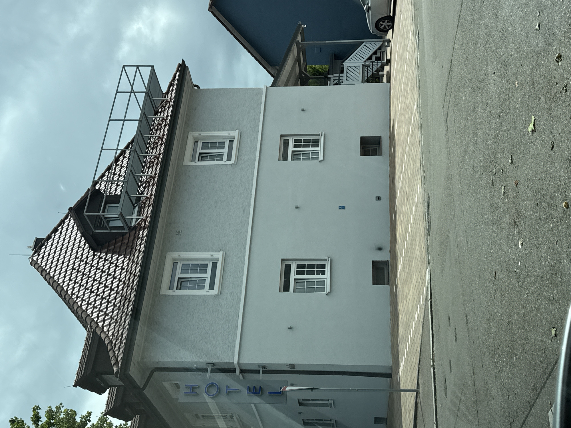 This image, captured in portrait orientation and rotated 90 degrees counter-clockwise, depicts a multi-story building, likely a hotel, situated next to a paved road under an overcast sky.
The building on the left features a traditional gabled roof with dark, textured tiles. A prominent metal framework, possibly for signage or an antenna, rises from the roofline. Below, the facade is a light-grey, textured stucco, with a white-framed, multi-pane window visible.
The central part of the building has a smoother, light-grey facade. Two more white-framed, multi-pane windows are present, alongside a smaller square window. At ground level, the reflection of the word "HOTEL" in blue, capital letters is clearly visible, confirming the building's purpose.
To the right of the main building, another structure with a dark blue roof and white outdoor stairs can be seen. A silver car is partially visible near this structure. The foreground and right side of the image show a paved surface comprising dark asphalt and a lighter section, possibly cobblestone. The sky is a uniform light grey, indicating an overcast day. Green foliage from trees is visible in the bottom left corner. The scene captures a street view in Tiengen, Germany.