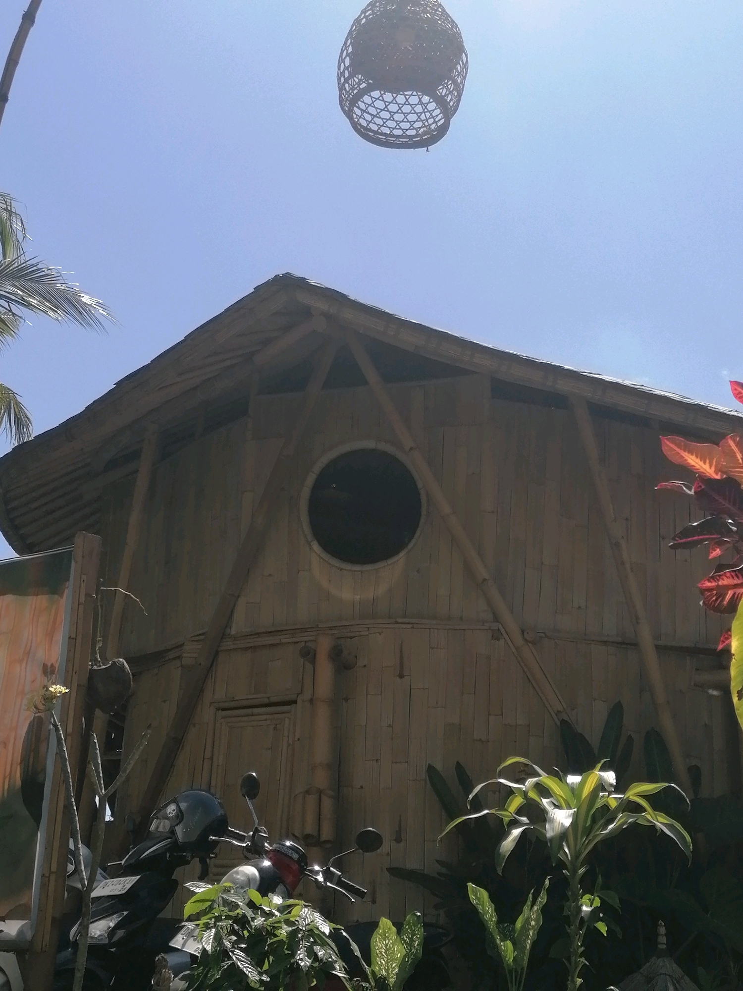 A wide, low-angle shot captures a traditional bamboo structure with a thatched roof against a clear blue sky in Ubud District, Indonesia. A circular window is visible on the side of the building. In the foreground, two motorcycles are parked, one with a dark helmet on its handlebars. Lush green foliage and plants surround the base of the structure and the motorcycles. A woven light fixture hangs from a bamboo pole in the upper right portion of the frame. The scene appears to be an outdoor area, possibly a guesthouse or a communal space, with a distinctly tropical ambiance.