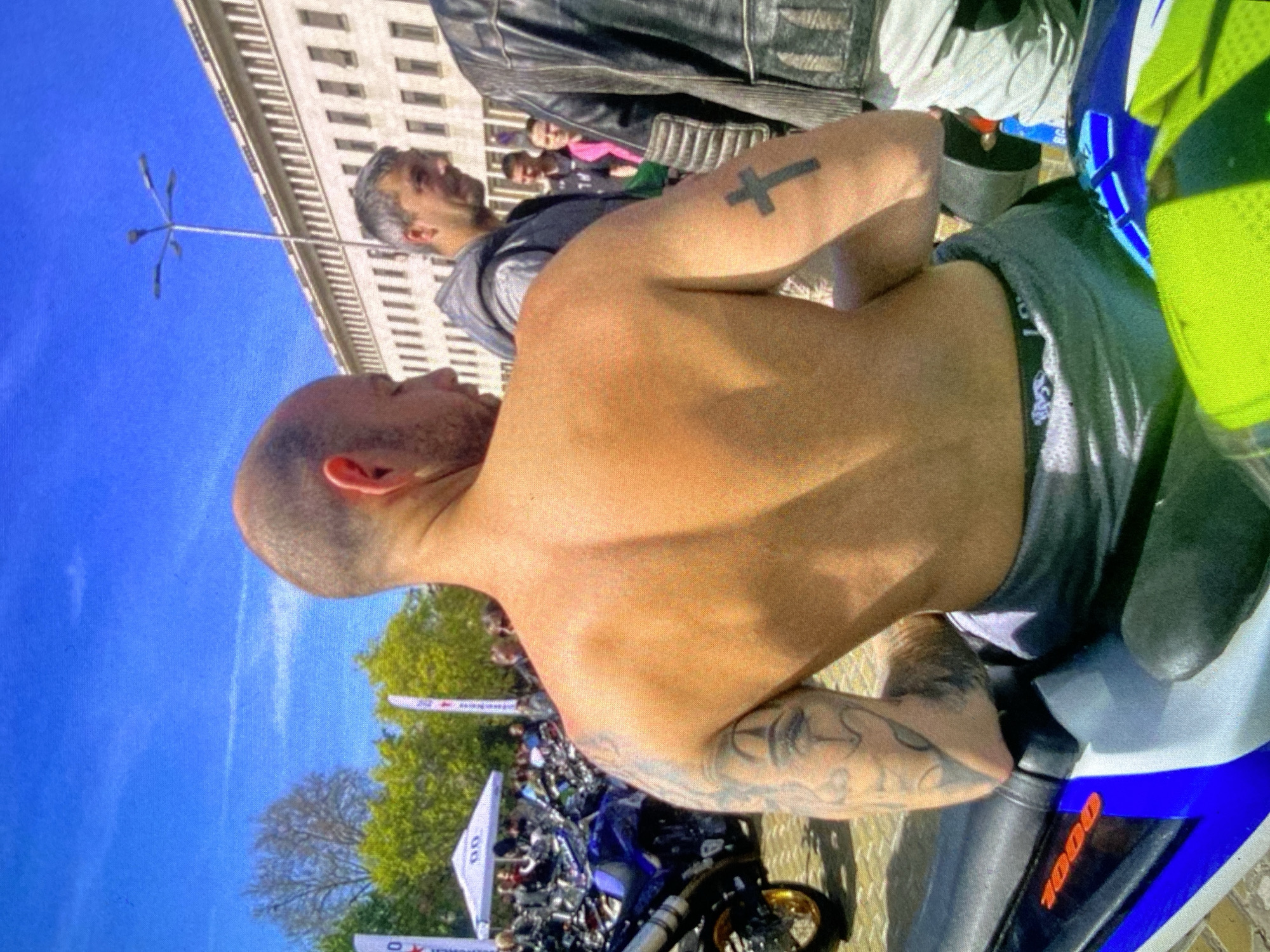 The image captures an outdoor scene, likely a motorcycle gathering or event in Bankya, Bulgaria, under a clear, bright blue sky during the daytime.
The primary subject is a man seen from the back and side, with a shaved head and a muscular physique. He appears to be shirtless or wearing a very low-cut vest, revealing his back and shoulders. A prominent black cross tattoo is visible on his left bicep, and his right forearm is adorned with intricate tribal or abstract tattoos. He is seated on a motorcycle that is predominantly blue, white, and black, featuring a gold-colored front wheel rim. The number "1000" is displayed in orange text on the side of the motorcycle. The man's underwear waistband, partially visible above his trousers, shows text that begins with "ROYAL...".
In the midground, another man with gray hair and a beard is visible, wearing a dark hoodie and a leather jacket. Several other blurred figures are scattered further back, suggesting a crowd. The background features a tall, light-colored building with multiple windows, typical of urban architecture, along with green trees. A unique street lamp or decorative pole with several radiating arms stands against the sky. Partially visible white banners or flags, some bearing the text "00" and possibly other branding, are present among parked motorcycles in the distance, reinforcing the context of a public event or rally.