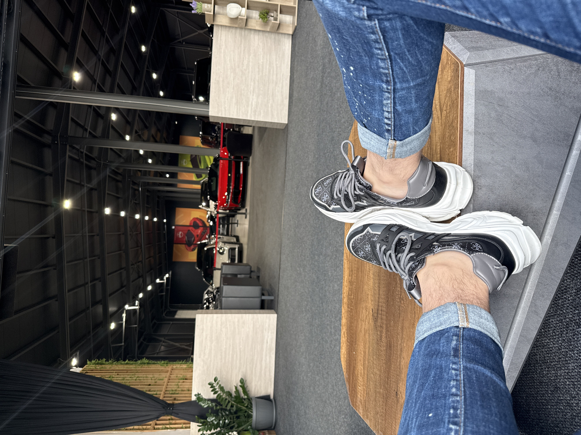The image, oriented vertically, captures a first-person perspective looking down at a person's feet and out into a large indoor space. In the foreground, the individual wears blue denim jeans, cuffed at the ankles, revealing light-colored skin and ankle hair. They are wearing a pair of black and grey athletic shoes with thick, sculpted white soles. The black sections of the shoes feature a repeating "G" logo pattern, characteristic of the brand Guess. The feet are resting on a smooth wooden surface, likely part of a table or bench, next to a dark grey, textured surface.
The background reveals a spacious, high-ceilinged indoor area, likely a car showroom or exhibition hall, possibly located in Plovdiv, Bulgaria. The ceiling is dark and features a prominent grid of metal beams, sparsely lit by numerous small, bright artificial lights. Several cars are on display within the hall, including a prominent red vehicle (possibly a sports car or SUV) positioned towards the right side of the frame. Other vehicles are visible further back. Various light-colored rectangular structures, serving as display pedestals or room dividers, are scattered throughout the space. One of these structures holds small decorative plants. A dark, pleated fabric drape and additional potted plants are visible in the lower left section of the frame, contributing to the modern, curated environment. The scene depicts a relaxed moment of observation within this commercial or exhibition setting.