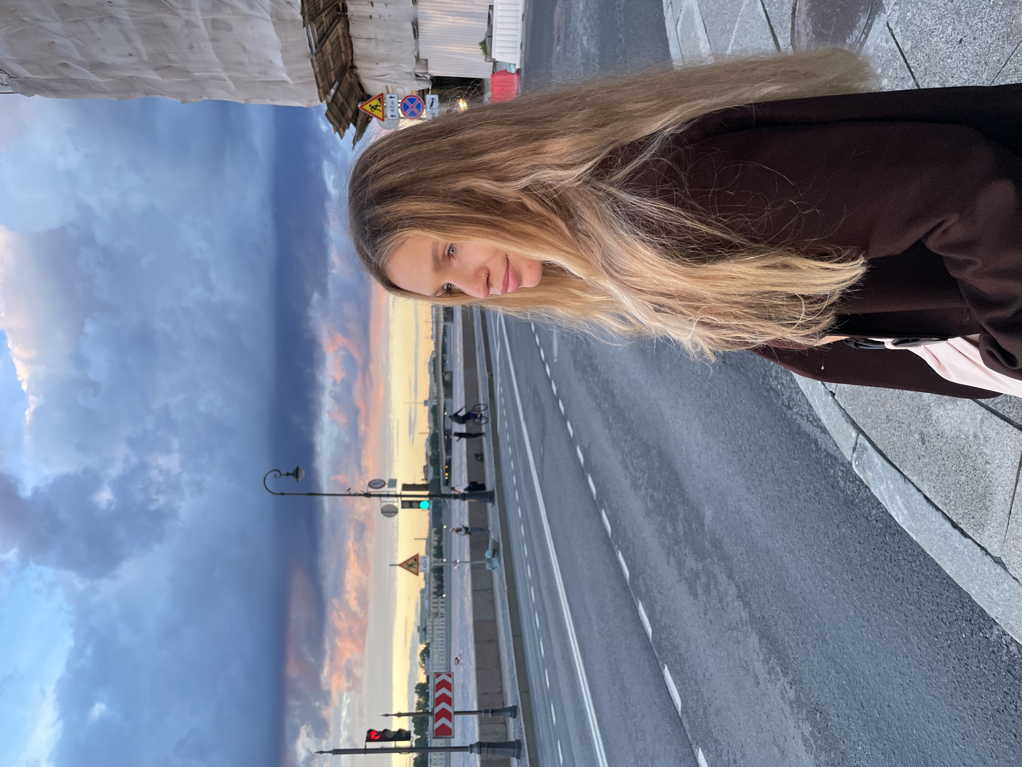 A young woman with long blonde hair is pictured in an outdoor setting. She is looking to her left, with a slight smile on her face, and appears to be posing for the photo. She is wearing a dark brown jacket over a light-colored shirt. The background shows a dramatic sky with clouds tinged with pink and orange, suggesting it is either sunrise or sunset. A road with white lane markings runs alongside a waterfront. Several traffic lights and streetlights are visible. There are also a few people in the distance, one of whom is riding a bicycle. The overall mood of the image is peaceful, with the soft colors of the sky creating a serene atmosphere. There are no visible signs with text.
