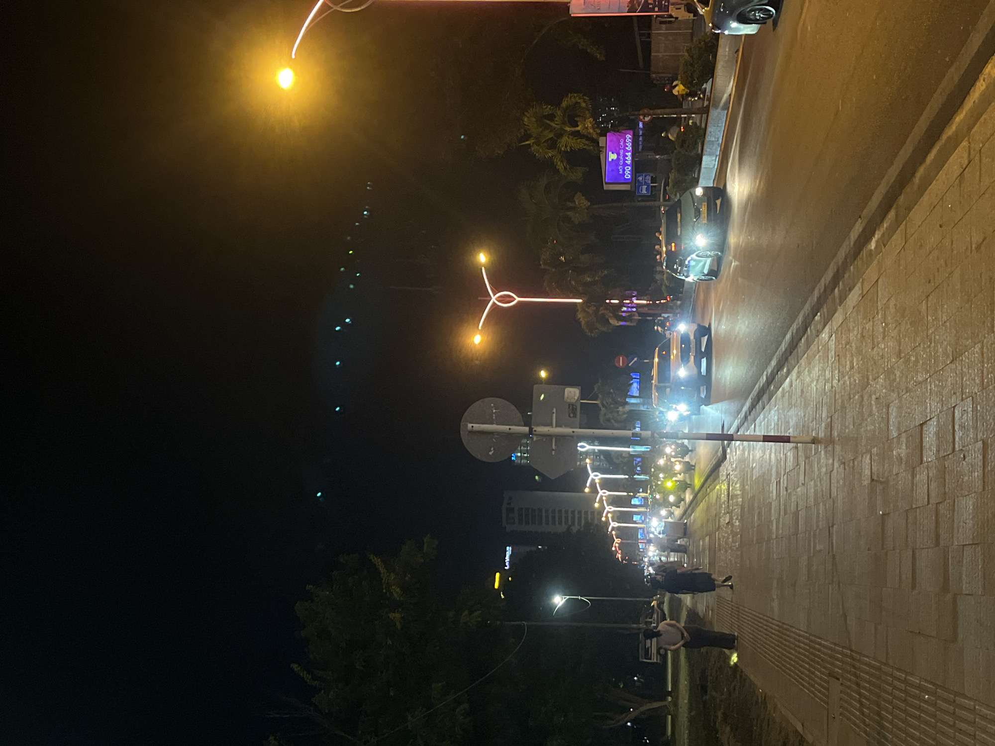The image depicts a night scene on a city street in Nha Trang, Vietnam, likely along a coastal road given the presence of palm trees and warm lighting. The perspective is from a sidewalk looking out at a street with vehicular traffic.
In the foreground, a paved sidewalk is visible, with textured paving stones indicating a pedestrian walkway. Two individuals are walking away from the camera on the sidewalk. The person on the left is wearing a light-colored top and dark pants, while the person on the right is wearing a dark dress.
The street is moderately busy with traffic, including cars and numerous motorcycles. Headlights and taillights illuminate the road. A white car is in the left lane, and a green car is in the right lane, both with their headlights on. A dark-colored car is partially visible in the far right lane. A traffic light pole with a red and white striped base stands on the sidewalk.
The background shows a typical urban night environment. Streetlights cast a warm, yellow glow. Decorative lights, possibly part of a festive display or permanent street decoration, are visible along the median of the road, forming an illuminated archway pattern. Beyond the street, there are buildings, including a tall, modern high-rise building with illuminated windows. Palm trees are scattered throughout the scene, suggesting a tropical or subtropical location. A large, purple digital billboard with Vietnamese text and a phone number is mounted on a pole. The sky is dark, with a few distant green lights visible. The overall impression is a lively street scene at night in a Vietnamese city.