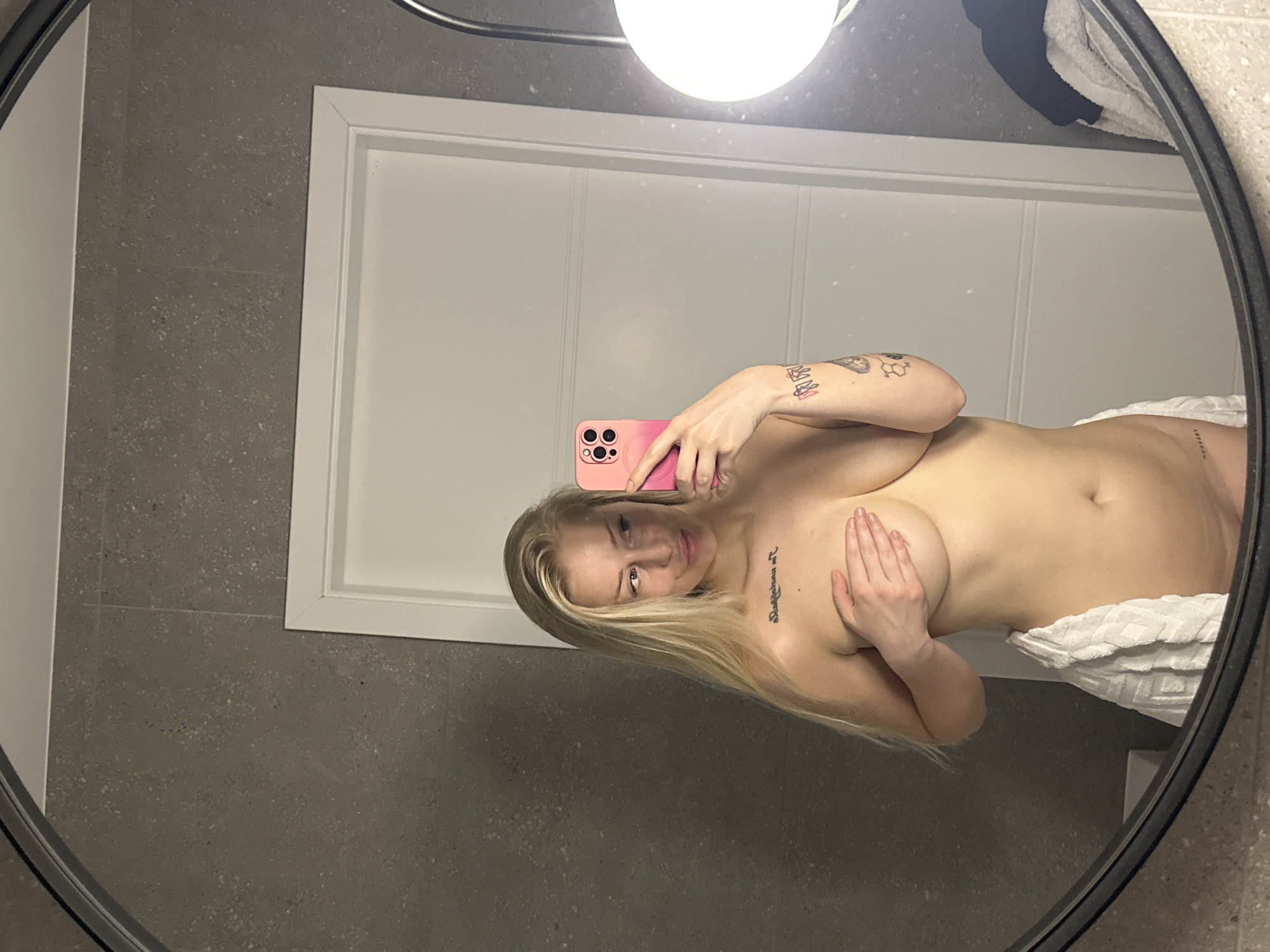 The media file shows a woman taking a mirror selfie. She has blonde hair and is holding a pink phone. Her upper body is partially visible, and she is wearing a white towel around her lower body. There are tattoos on her arms and torso. The setting appears to be a bathroom with grey tiled walls and a white door in the background. The lighting suggests it is indoors, possibly during the day. The woman's expression is neutral. The image is a reflection in a round mirror.