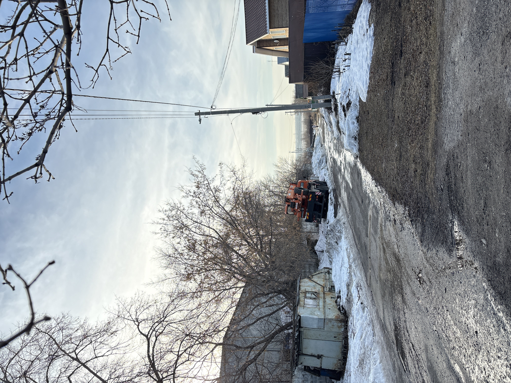 This image depicts a semi-rural street scene in Ulyanovsk, Russia, during what appears to be late winter or early spring. The ground is characterized by a muddy, unpaved or severely degraded asphalt road, saturated with moisture and featuring puddles, indicating melting snow. Dirty snow patches persist along both sides of the road.
On the left side of the road, a dilapidated, rusted light-blue metal structure, resembling a utility shed or small trailer, is partially buried in snow and debris. Behind this structure, a large, utilitarian building with light-colored, possibly corrugated metal siding is visible. An orange and black heavy-duty truck, consistent with a specialized utility or crane vehicle, is parked near the dilapidated structure. Bare deciduous trees with intricate branch networks line this side of the road, obscuring parts of the background. A dark-clad figure is partially visible near the left edge of the dilapidated metal structure.
On the right side of the road, a concrete utility pole stands prominently, supporting multiple power lines and a street lamp fixture. Further down this side, several modern-style residential houses with brown tiled roofs and light-colored facades are visible, separated from the road by a vibrant blue corrugated metal fence. Patches of bare ground with dried vegetation and melting snow are present in front of the fence. A solitary individual, dressed in light-colored outerwear, is observed walking away from the viewer on the left side of the utility pole.
The sky is partially overcast, with brighter areas indicating diffused sunlight. A faint, distant urban or industrial landscape can be discerned on the horizon in the background.