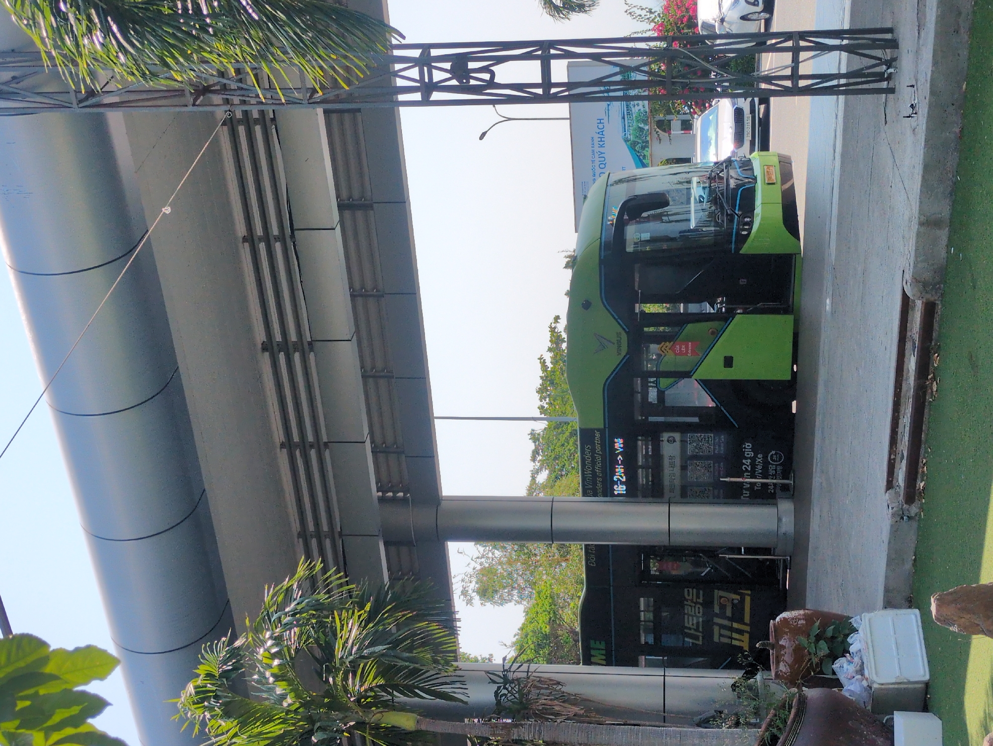 The image shows a green and black VinBus electric bus parked at a covered bus stop in what appears to be Nha Trang, Vietnam. The bus has the VinBus logo and various advertisements in Vietnamese and Korean. The bus stop structure is modern, with metal supports and a covered roof. Tropical foliage is visible around the base of the supports. A sign reads "NG QUỐC TẾ CAM RANH - QUÝ KHÁCH" which translates to "Cam Ranh International Airport - Welcome." Potted plants and discarded containers sit near the bus. A portion of a grassy area is visible. Other cars are parked across the street.