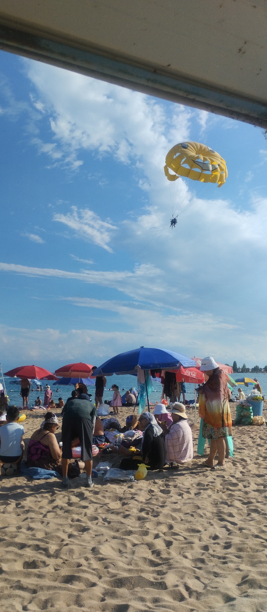 The image captures a busy beach scene in Cholpon-Ata, Kyrgyzstan, on a clear day with scattered clouds. In the sky, a yellow parasail shaped like a smiley face is visible, with two individuals harnessed beneath it, appearing to be parasailing. The beach itself is populated with numerous people relaxing under various beach umbrellas, including red, blue, and multi-colored ones. Several individuals are seated on the sand, some appearing to be in groups, with beach belongings like bags and towels spread around them. In the background, the calm water of Issyk-Kul Lake is visible, with more people wading or swimming near the shore. The sand is a light brown color, showing the footprints and texture of a well-used beach. The overall impression is one of a lively summer day at a popular tourist destination.