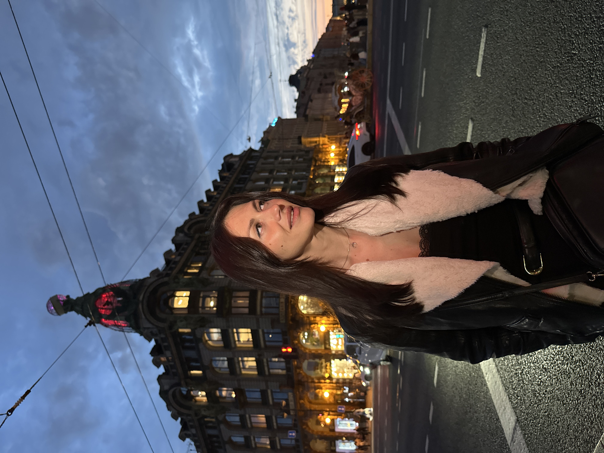 This is a medium shot photograph taken outdoors in Saint Petersburg, Russia, at dusk. The main subject is a young woman with long, dark hair, looking to her right. She is wearing a black leather jacket with a white shearling lining over a black top, and a necklace with a crescent moon pendant. A black crossbody bag is slung over her shoulder.
In the background, a grand, ornate building with illuminated windows and a distinctive dome topped with a globe-like structure is visible. The building's architecture suggests a historical significance, consistent with the city's aesthetic. The sky above is a deep blue with dramatic clouds, characteristic of twilight.
The scene is set on a city street, with visible road markings and streetlights casting a warm glow. Traffic lights are visible in the distance. Several vehicles, including a white car and a horse-drawn carriage, are present on the street. Pedestrians are also scattered along the sidewalk, and more buildings line the street stretching into the distance. Electrical wires are strung across the sky, further indicating an urban setting. The overall atmosphere is that of a vibrant city evening.