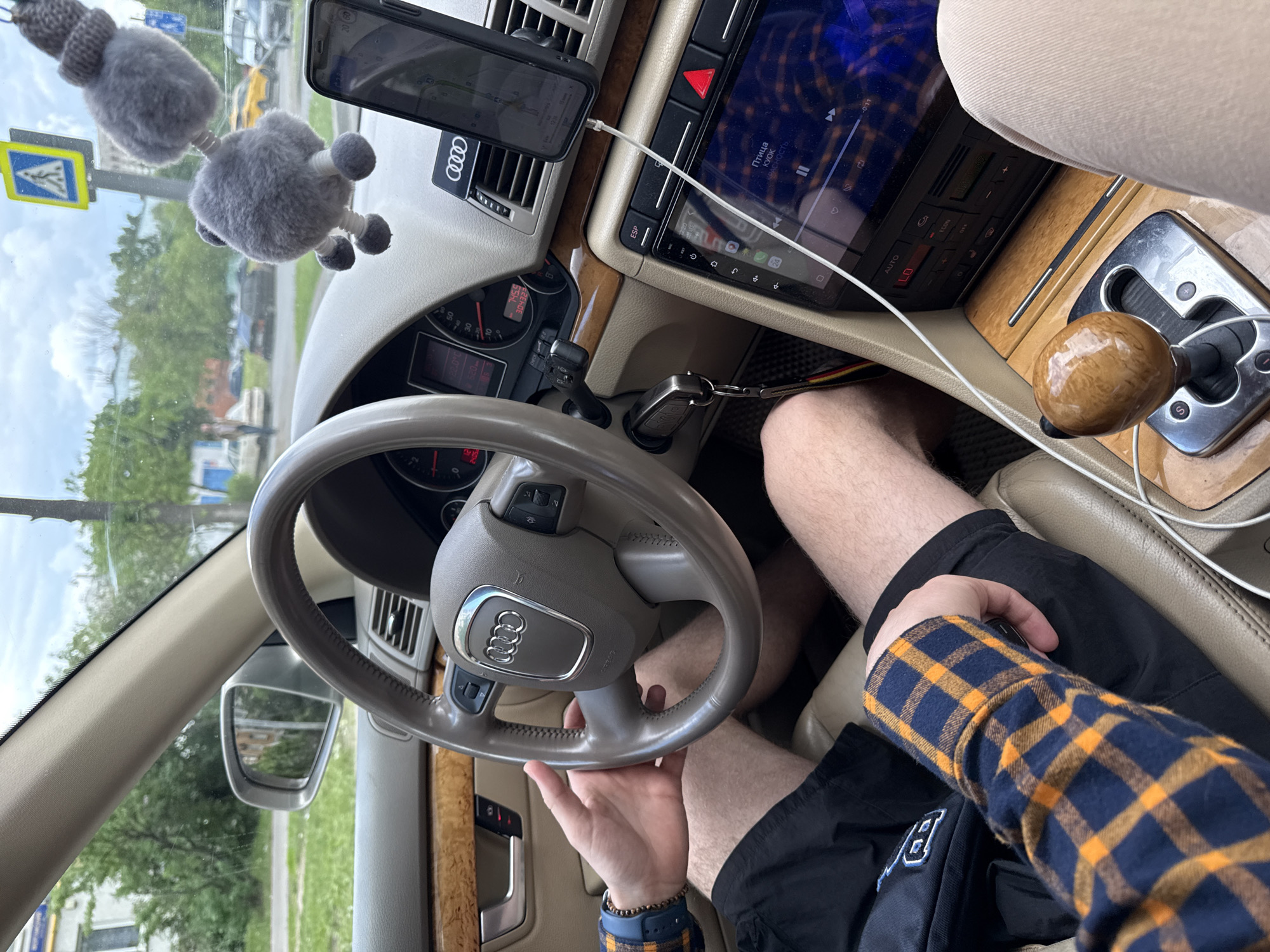 A photograph taken from inside an Audi sedan shows a person holding the steering wheel, with a smartphone attached to the dashboard via a vent mount displaying a navigation app. The car's entertainment system is also visible, showing a music player interface with Russian text. A fuzzy grey car decoration hangs from the rearview mirror. The driver is wearing a blue and orange plaid long-sleeve shirt and black shorts. The dashboard and interior are beige, and a wooden-handled gear shift knob can be seen to the right of the driver's leg. The key is in the ignition. A pedestrian crossing sign is visible through the windshield, as well as a street view, which may indicate a location in Moscow, Russia.