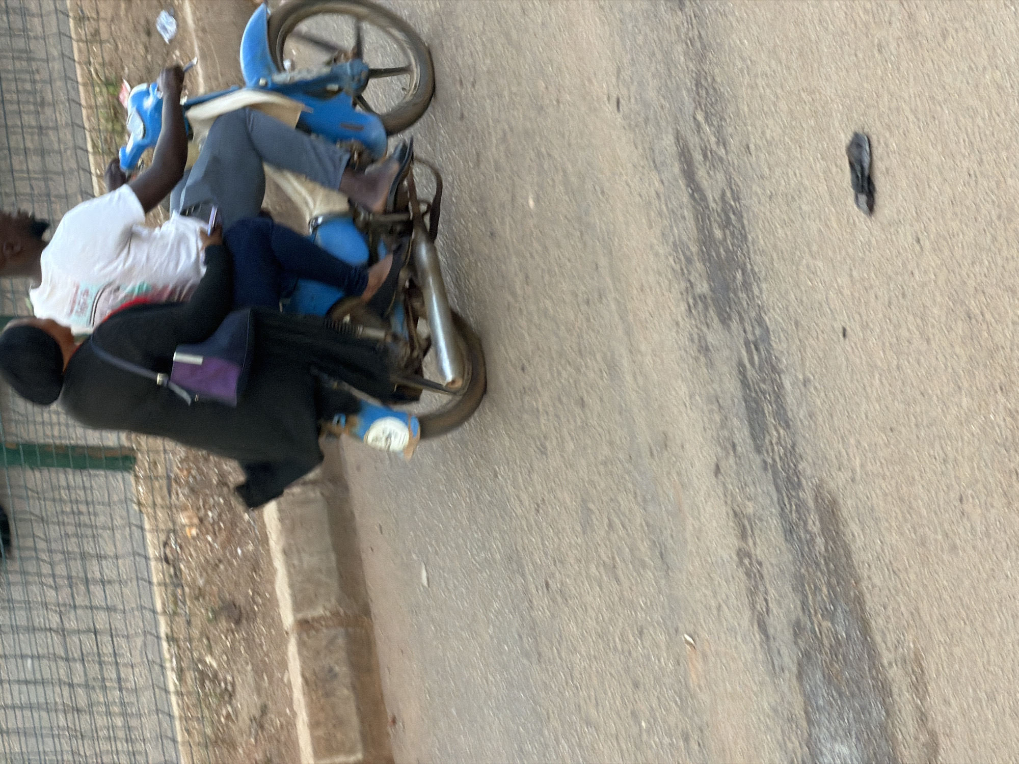 Two individuals are riding on a blue motorcycle. The person in the front, wearing a white t-shirt and grey pants, is operating the motorcycle. The person in the back, wearing a black coat and dark blue leggings, is holding onto the operator. They are on a paved road in Bukuru, Nigeria. The environment appears to be an urban or semi-urban setting with a fence and a concrete barrier visible in the background. The overall scene is captured in daylight.