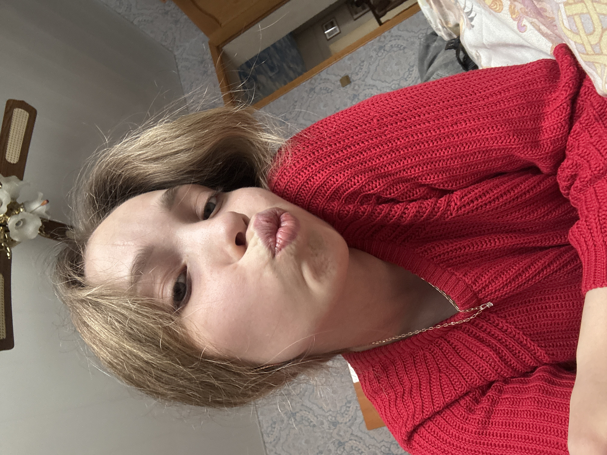 A fair-skinned person with light brown hair wearing a red knitted sweater and a thin gold necklace is posing for a selfie. The person is making a kissing face with their lips pursed. The background shows a bedroom setting with patterned wallpaper, a bed with patterned sheets, and a wooden headboard or door. The location appears to be an interior setting, potentially a home. The city of Прикамский, Russia, cannot be determined from this image.