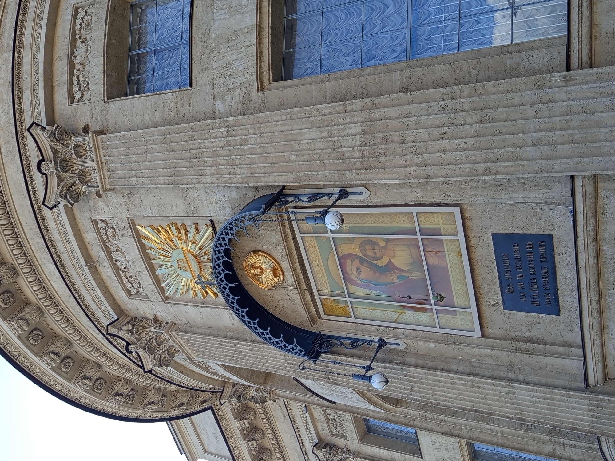 This is an exterior, low-angle shot of a building's facade, likely taken during the daytime under clear skies. The architecture is classical and ornate, featuring a large, curved section with intricate carvings and decorative elements. A prominent fluted column is visible on the right side of the image, extending upwards.
Mounted on the building is a dark, curved metal sign bracket with decorative scrollwork. Hanging from the bracket is a single globe-shaped light fixture. Above the bracket, a stained-glass window depicts a religious icon of the Virgin Mary holding the Christ child, rendered in muted colors. To the left of the stained-glass window, a golden sunburst motif with a cross at its center is mounted.
Further to the right, a dark rectangular plaque with Cyrillic text is affixed to the wall. The text, though partially obscured, appears to be in Russian. The building's facade is constructed from light-colored stone, possibly limestone or sandstone, with a textured finish. Several horizontal, stepped architectural elements are visible, creating a layered effect. The overall impression is one of historical and religious significance, consistent with a church or a similar institution in Saint Petersburg, Russia.