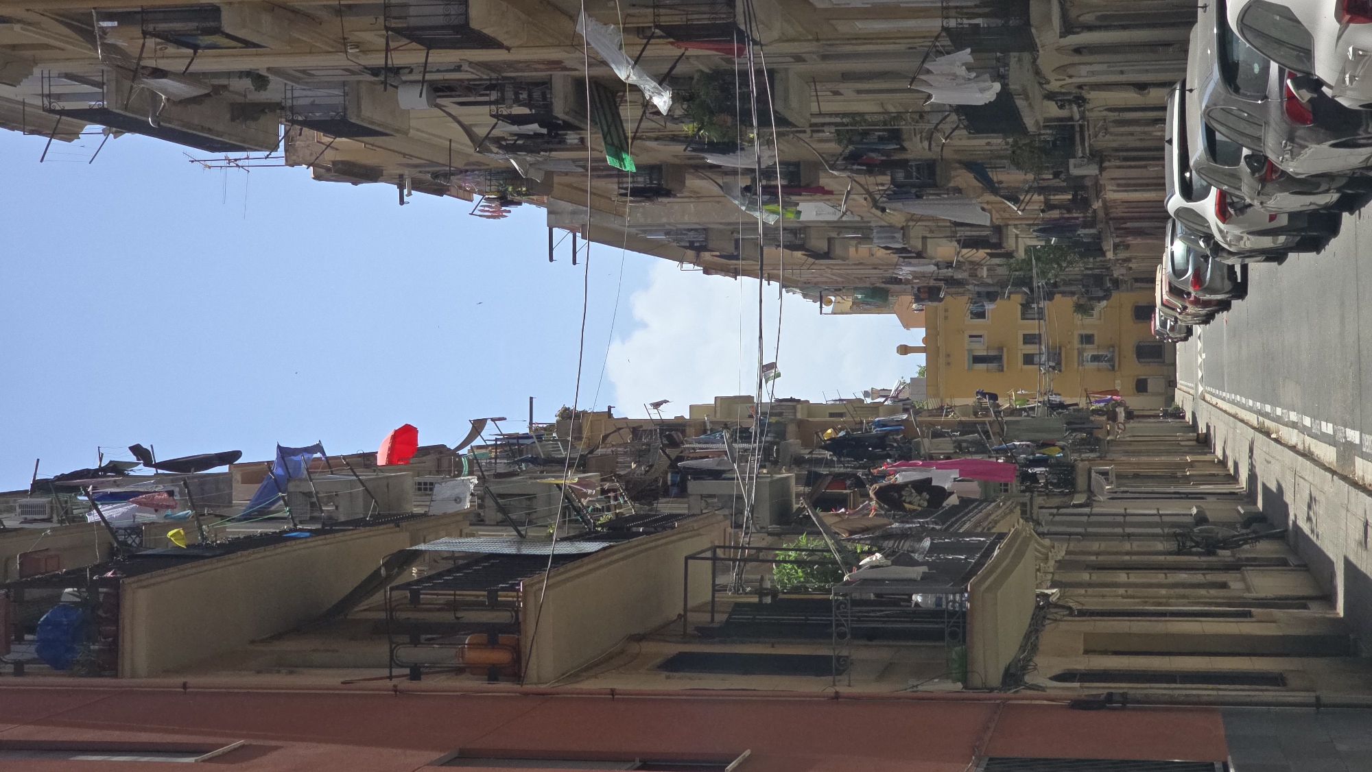 This image depicts a street scene in Barcelona, Spain, with buildings on either side. The perspective is from a high vantage point looking downwards, and the image is rotated 90 degrees clockwise.
On the right side of the image, a row of cars is parked along a paved street, with a sidewalk bordering it. The cars are of various colors, including silver and white. The street appears to be an urban environment.
The left side of the image is dominated by the facades of multi-story buildings. These buildings have an aged appearance, with many balconies and windows. Laundry is draped over many of the balconies, suggesting it is a sunny day with clothes being air-dried. The balconies are cluttered with various objects, including tarps, storage containers, and other household items, giving a sense of lived-in, perhaps crowded, urban living. A bright red umbrella or similar object is visible on one of the balconies. A yellow building stands out among the others.
The sky is a clear blue with a few scattered white clouds, indicating fair weather. The lighting suggests it is daytime, likely midday or early afternoon. There is no visible text in the image. The overall impression is one of a vibrant, densely populated urban area with everyday life unfolding on its streets and balconies.