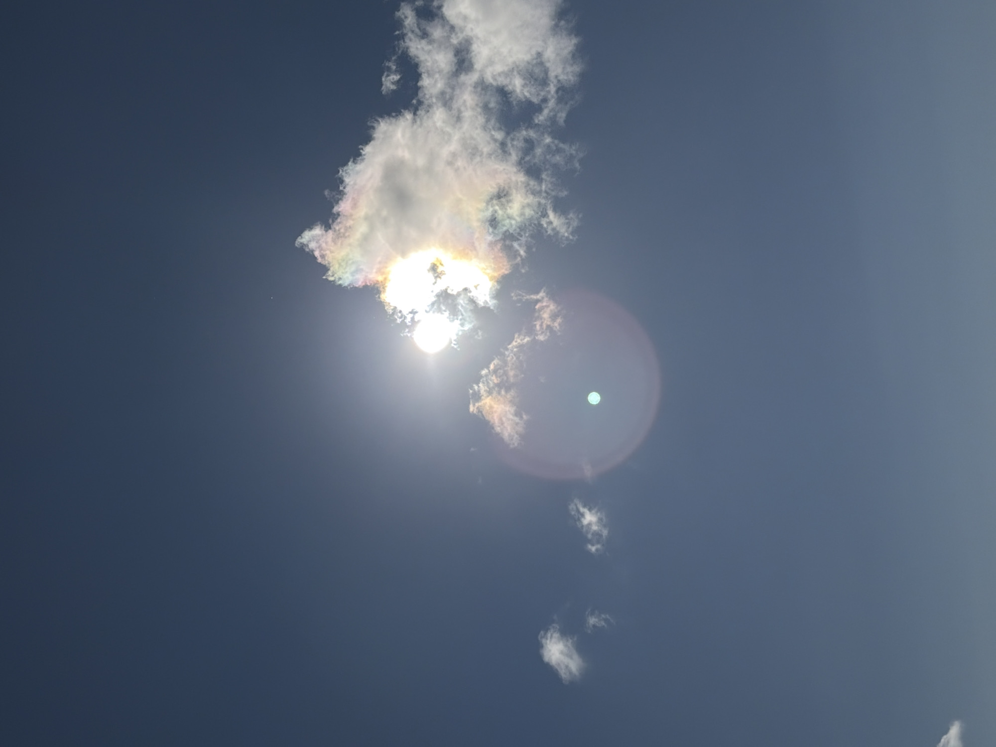 The image shows a bright sun partially obscured by a cloud with iridescent colors, indicating a sun halo or iridescence. A faint, circular lens flare is visible in the lower center of the frame. There are also two small clouds to the left of the sun. The sky is a clear, deep blue. The image is a photograph of the sky taken outdoors. There is no information in the image to suggest it is located in the city of Ozerskoe, Russia, or any other specific location. There are no people, objects, or actions identifiable in the image other than the celestial phenomena and clouds.
