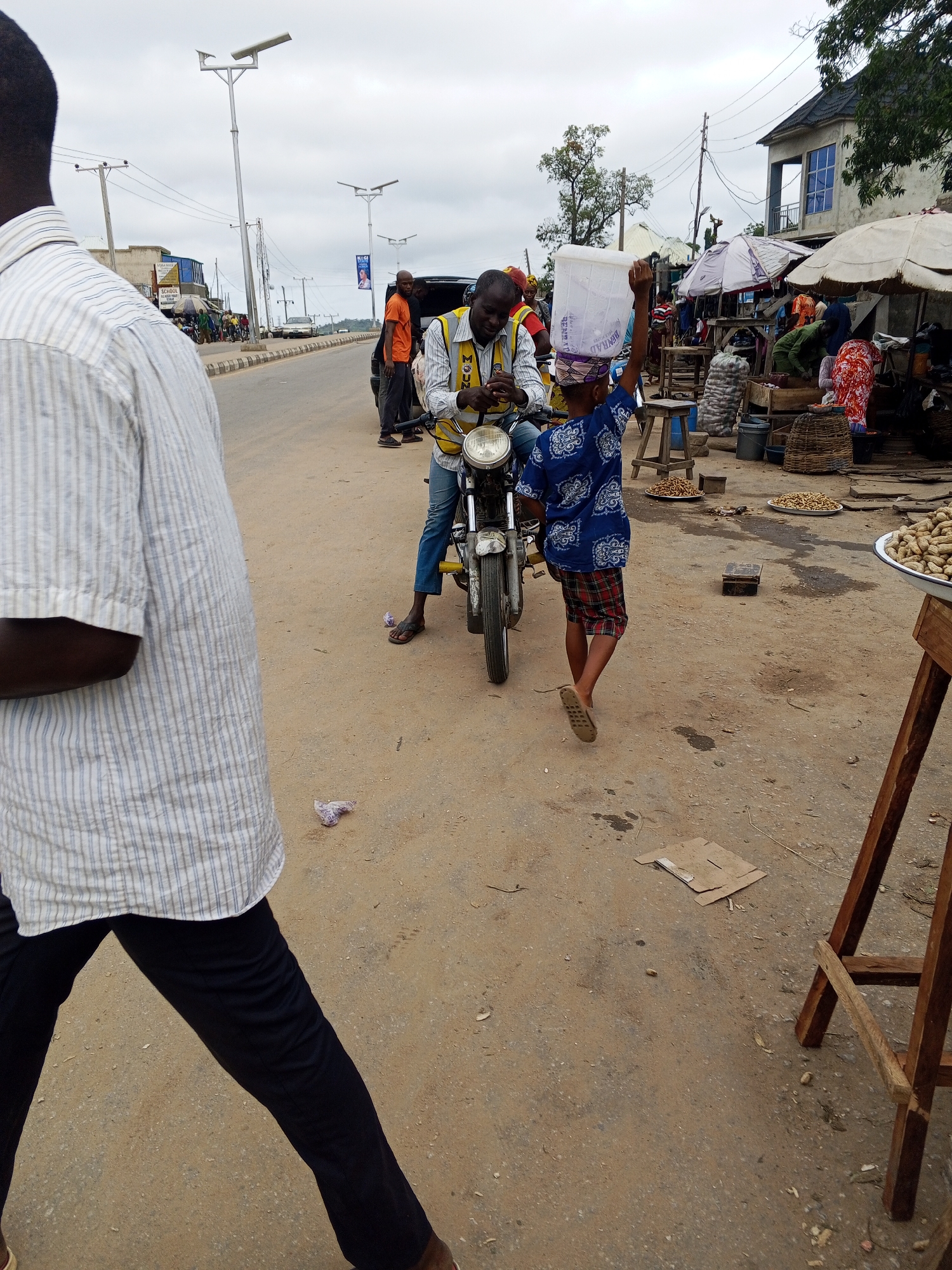 In Zango-ndaji, Nigeria, a street scene unfolds with a man in a striped shirt and black pants walking away from the camera towards the left foreground. To his right, a man wearing a yellow vest over a white shirt and blue jeans sits on a motorcycle. He is looking towards the right. A young person, with their back to the camera, walks away from the motorcycle towards the right midground, carrying a large white plastic container on their head. This individual wears a blue patterned shirt and red plaid shorts. The background shows a street with other people, market stalls with umbrellas, buildings, and utility poles. The ground is dusty and uneven, with scattered debris.