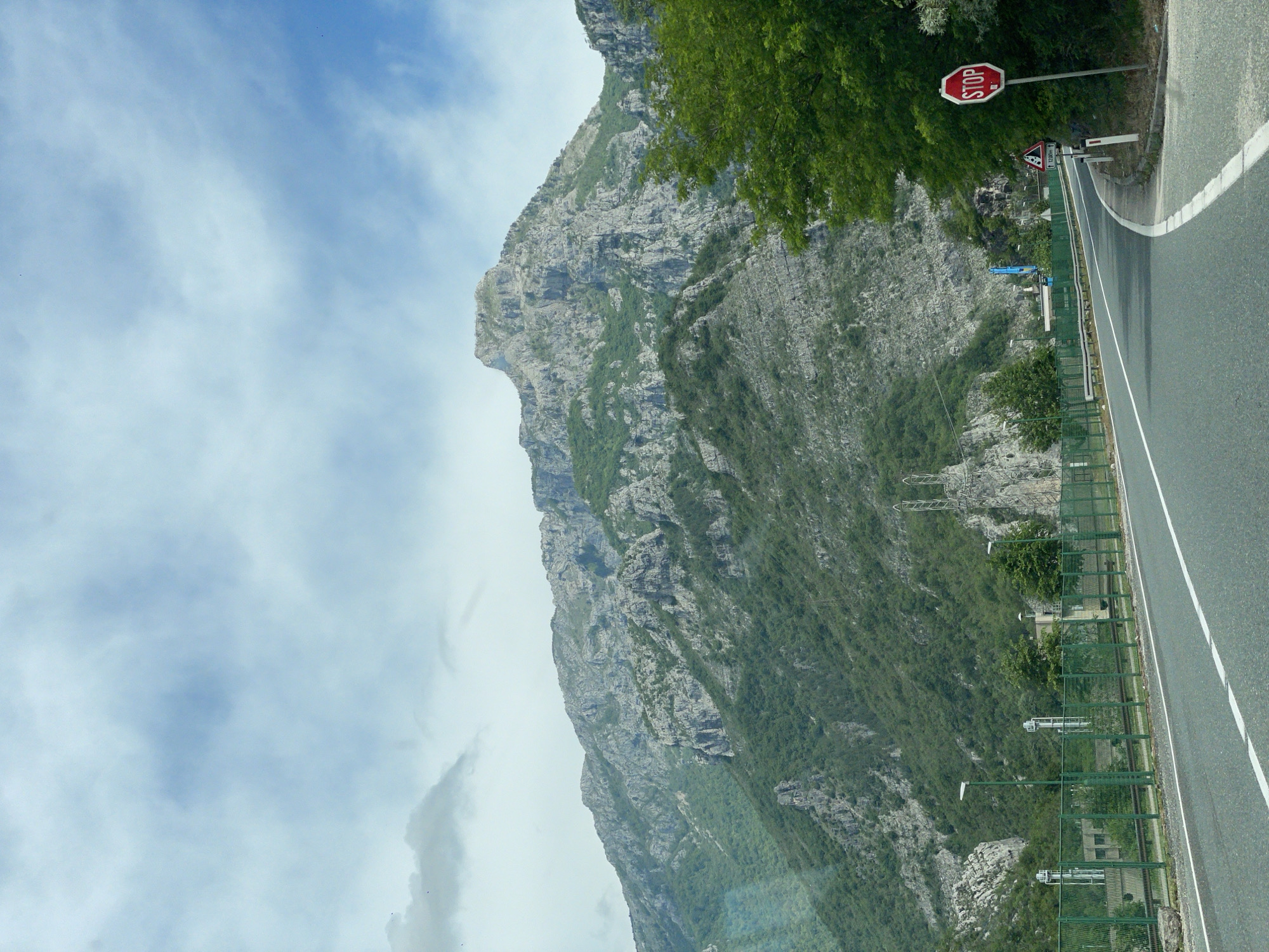 A medium-angle shot depicts a paved road running alongside a steep, rocky mountain covered in sparse greenery under a cloudy sky. A green guardrail borders the road, and a red stop sign is visible on the right side of the road near some trees. Power lines are also present on the mountainside. The location is Donji Jasenjani, Bosnia and Herzegovina.
