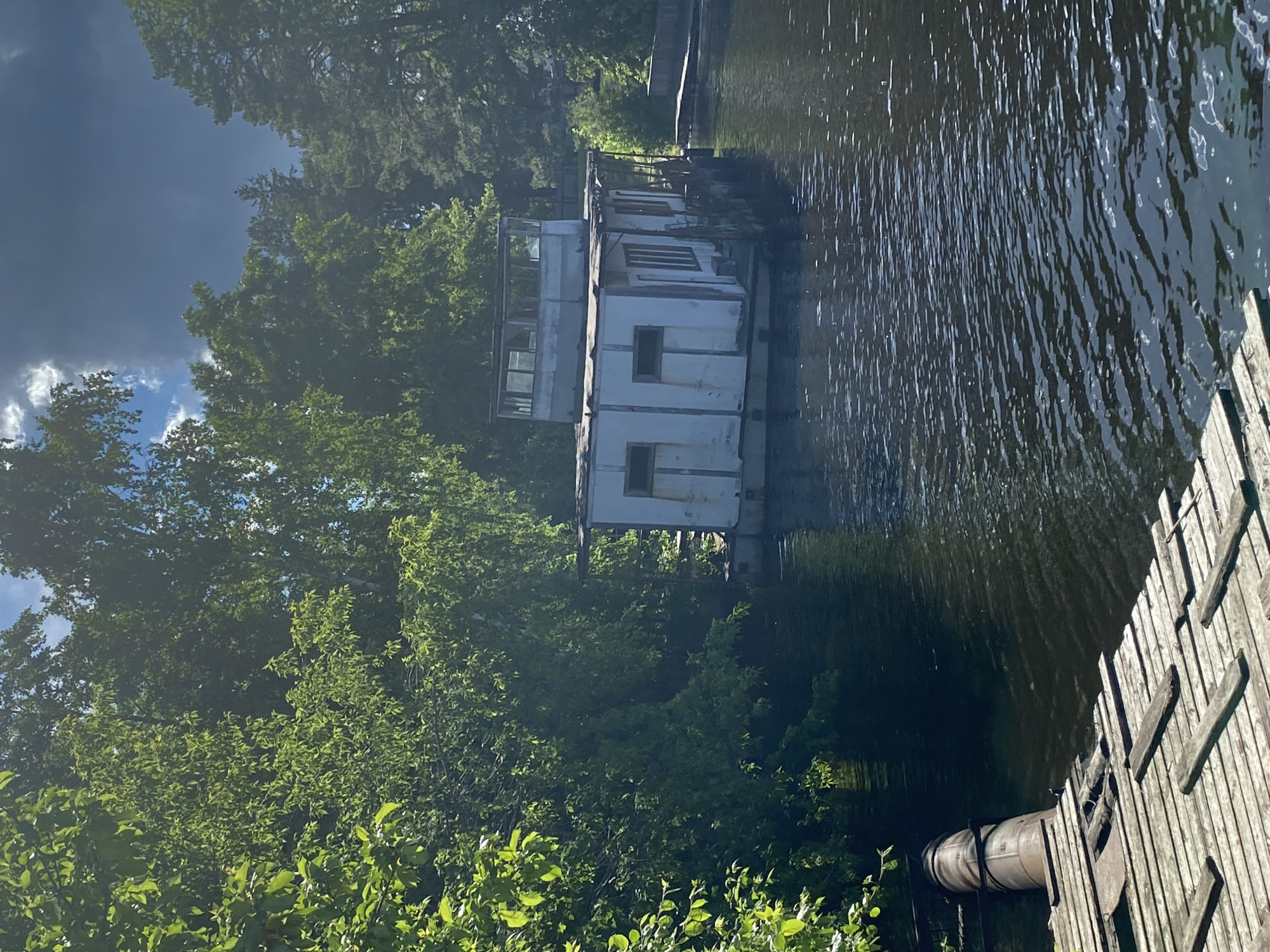 The image depicts a dilapidated houseboat on the Kama River in Chaikovsky, Russia. The houseboat is white and box-shaped, with visible windows and multiple levels. It appears weathered and in disrepair. The boat is moored alongside a wooden dock. A section of what appears to be a pipe is visible near the dock. The surrounding environment includes dense green trees. The sky is partially obscured by trees, with a dark storm cloud visible. The water of the river is dark and slightly rippled.