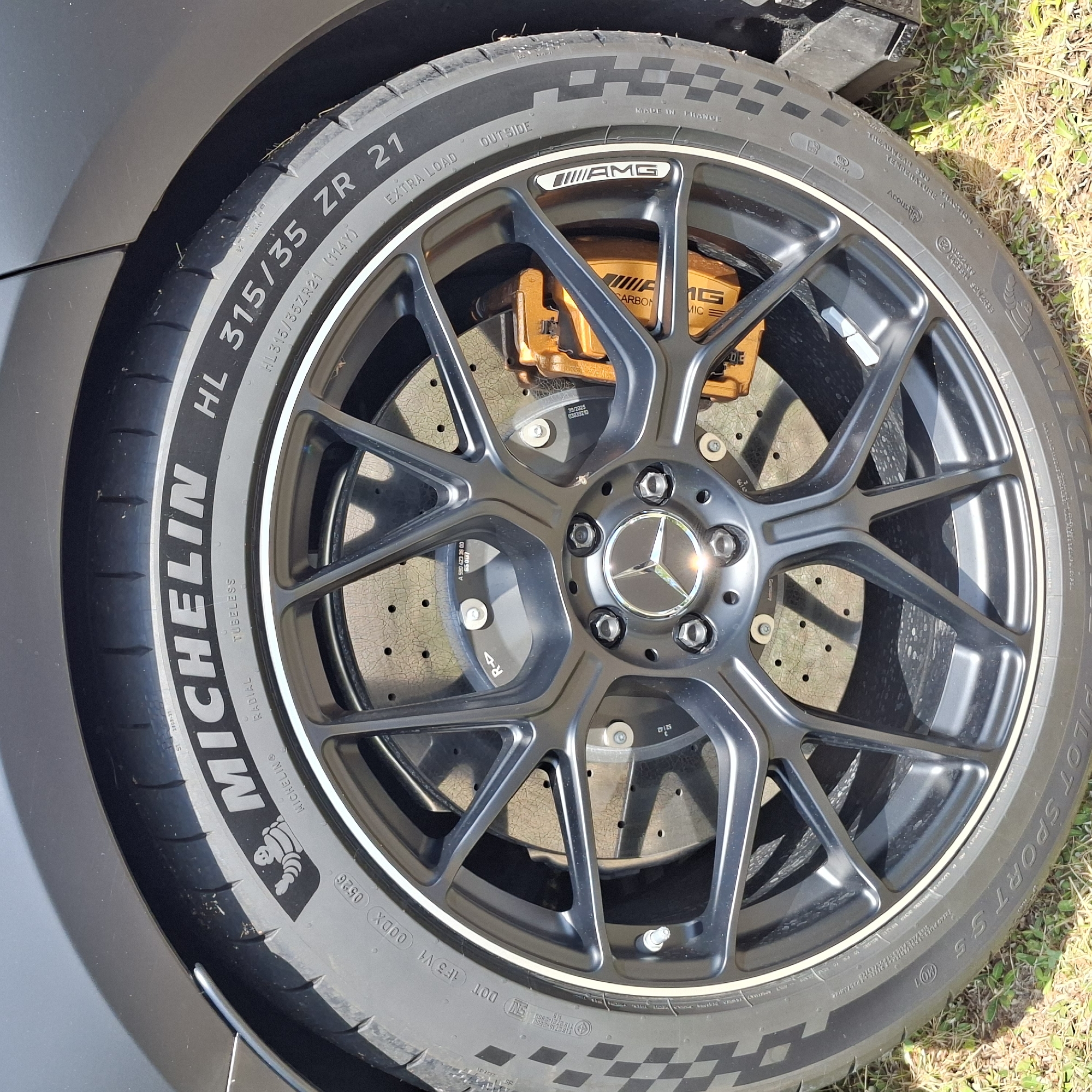 This media file is a close-up, eye-level shot of a car wheel, specifically a Michelin Pilot Sport tire mounted on a multi-spoke black AMG alloy wheel. The car appears to be dark gray. The wheel features bright orange brake calipers, also branded with AMG and "CARBON CERAMIC". The tire has prominent white lettering indicating its size "HL 315/35 ZR 21" and brand "MICHELIN". Additional text on the tire includes "MADE IN FRANCE", "EXTRA LOAD", "OUTSIDE", and various tire code markings. The wheel hub displays the Mercedes-Benz star logo. The car is parked outdoors on a grassy area, with some dried grass and sunlight visible on the ground and casting shadows on the wheel. The lighting suggests it is daytime, likely sunny. The setting appears to be a parking area or roadside given the presence of grass. No people are visible in the image. The focus is entirely on the automotive components, highlighting the tire, wheel, and braking system.