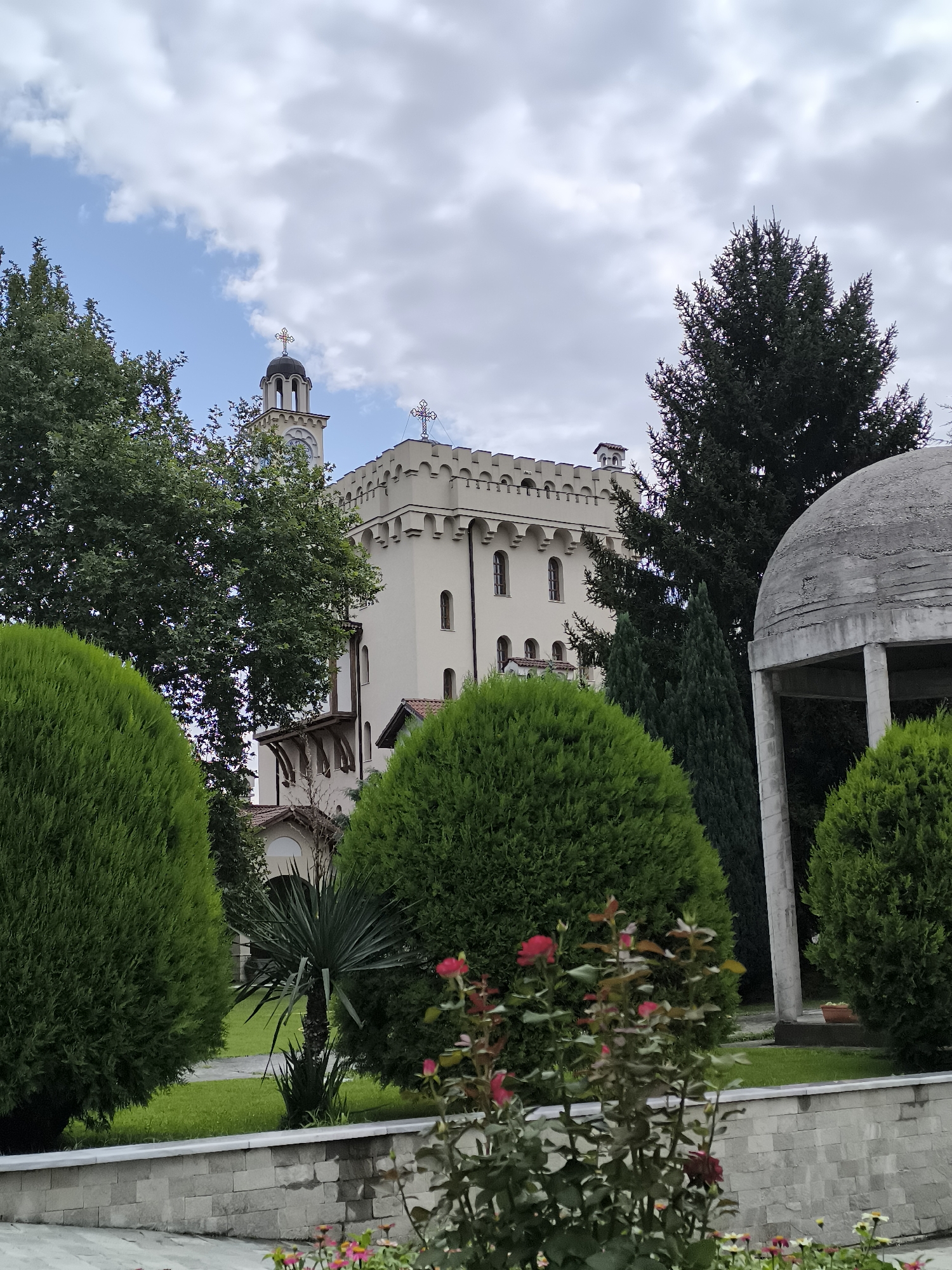 In Hadzhidimovo, Bulgaria, a cream-colored building resembling a castle or a fortified church stands prominently. Its top features a crenellated parapet, and a bell tower with a dome and cross rises on the left side of the structure. The building has multiple windows and a small turret. In front of the building, several manicured green shrubs are arranged, some of which are cone-shaped and others are rounded. A concrete dome-shaped structure with columns is partially visible on the right side of the image. In the foreground, a bed of roses is in bloom with several red and pink flowers. Lush green grass and more trees are visible in the background. The sky is partly cloudy with patches of blue visible.