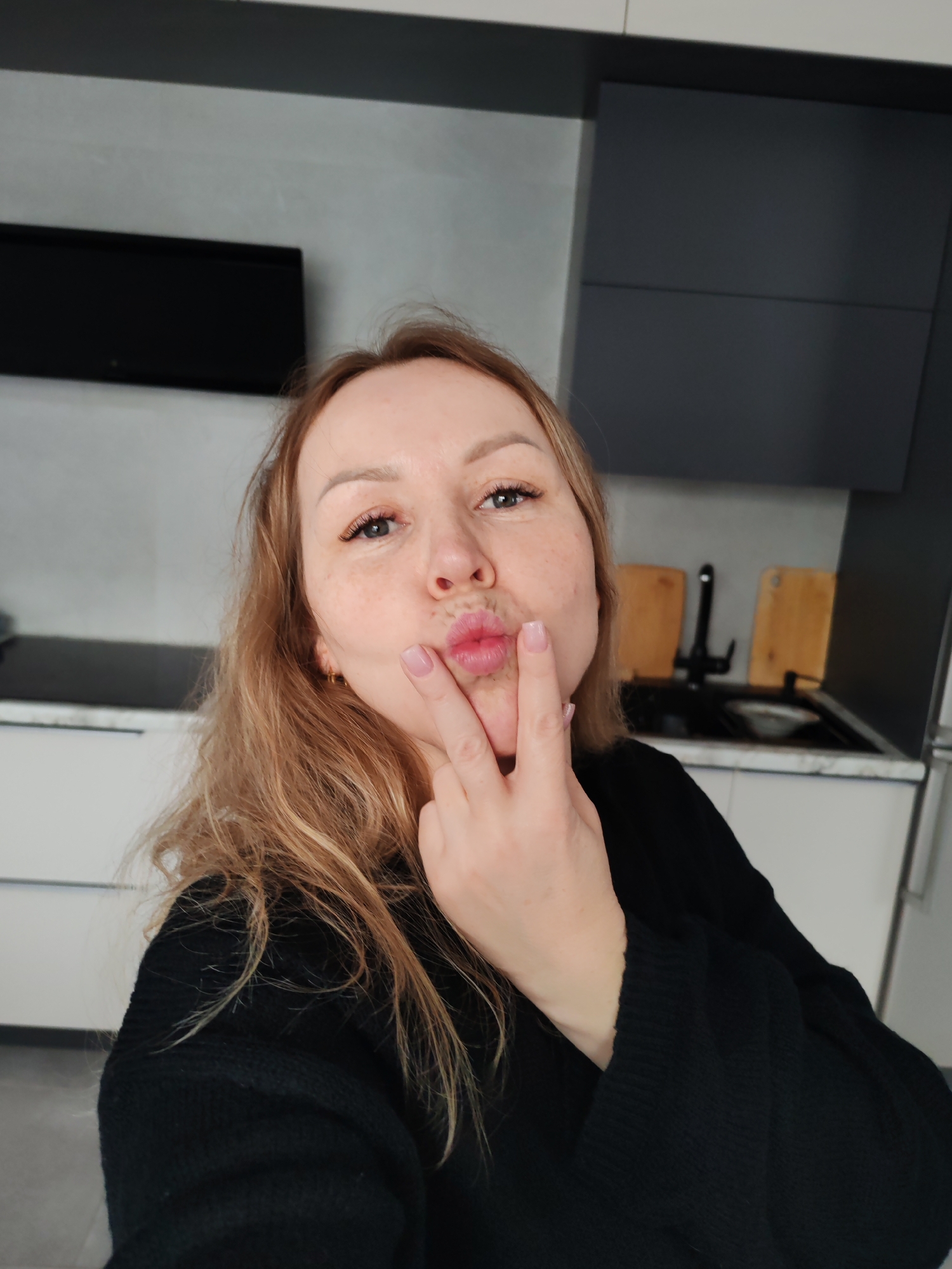 A woman is taking a selfie in what appears to be a kitchen. She has light skin, light brown hair, and is wearing a black sweater. She is puckering her lips into a "kiss" face and holding her index finger to her lips. The background features modern kitchen elements, including light-colored cabinets, a black countertop, and a black sink. A television is mounted on the wall. Although Chaikovsky, Russia, is provided, no information in the image confirms it to be that location.