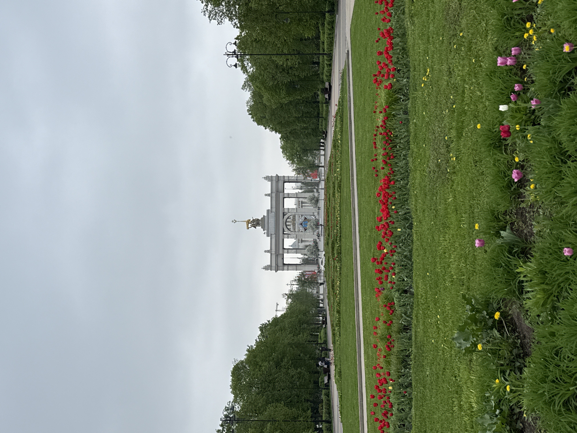 The image depicts the main entrance to VDNKh (Exhibition of Achievements of National Economy), a large exhibition and amusement park in Moscow, Russia. The prominent archway structure with sculptures at the top is the defining feature.
A paved pathway extends from the foreground towards the archway, flanked by lawns on either side. A line of red flowers borders the pathway on the right. Trees line the path on the left.
The sky is overcast. There are people walking in the park.