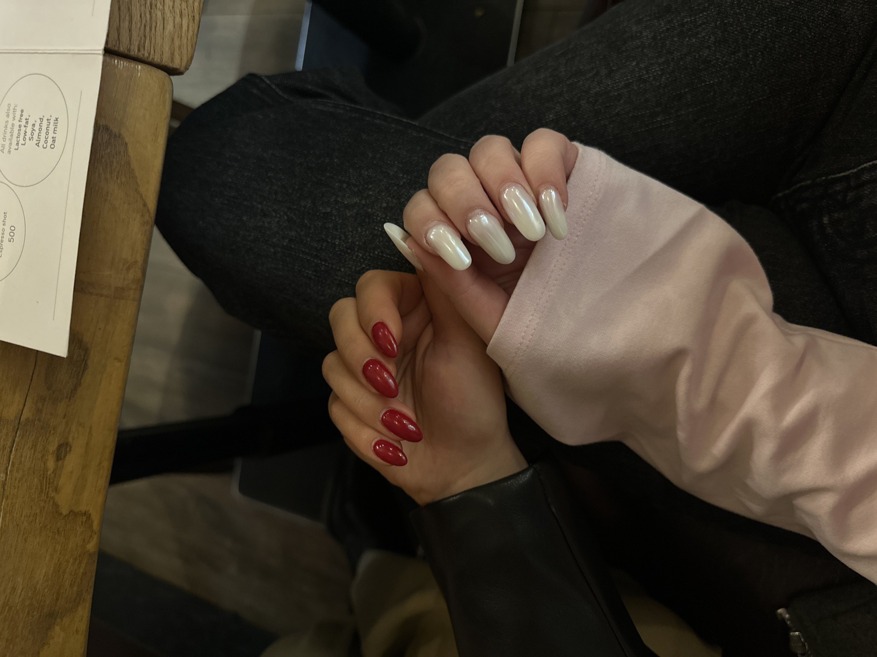 A pair of hands are shown interlocked on denim fabric. One hand, adorned with red nail polish, wears a black leather sleeve. The other hand, covered by a light pink sleeve, has pearlescent white nails. A wooden table surface and a menu listing drink options, including "Espresso shot 500" and various milk alternatives like "Lactose free," "Low-fat," "Soya," "Almond," "Coconut," and "Oat milk," appear at the top left.