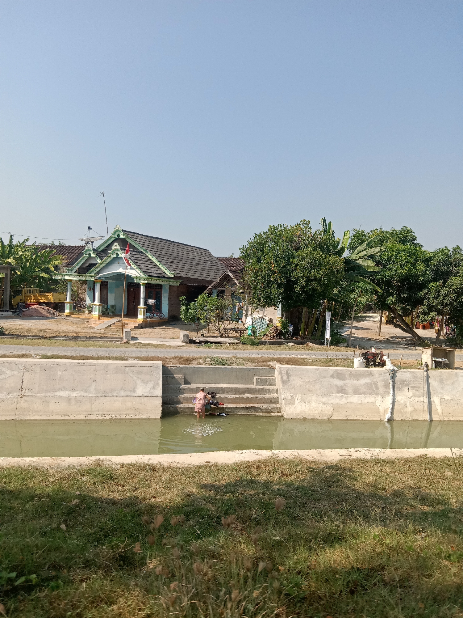 In Klepek, Indonesia, a rural scene unfolds under a clear blue sky. A canal with concrete retaining walls runs through the foreground, its murky green water reflecting the sky. On the stepped concrete edge of the canal, a person wearing a light-colored garment is partially submerged in the water, engaged in washing. Nearby, laundry is piled up. Behind the canal, a dirt path separates it from a cluster of traditional houses and trees. One house, with a green-trimmed roof and white pillars, stands prominently. Various trees, including banana trees, are visible, along with a small satellite dish mounted on a pole above one of the houses. To the left, a yellow truck and piles of sand suggest ongoing construction or activity. In the mid-ground, another person is visible near the house, and a bicycle is leaning against it. The overall impression is one of a tranquil, everyday life in a tropical, rural setting.