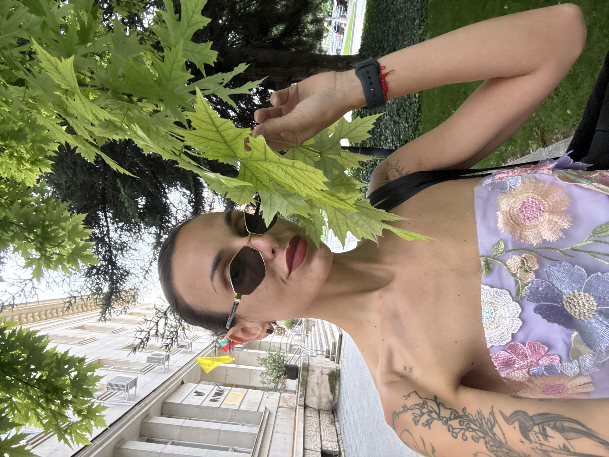 A woman with dark hair pulled back, wearing a strapless lavender top with floral embroidery, large sunglasses, and dark lipstick, holds a bright green leaf in her right hand. She stands outdoors, with a building and trees in the background. The building appears to be institutional, with windows and air conditioning units visible. A yellow flag with Cyrillic script and a Bulgarian tricolor flag are mounted on the building's facade. The woman has tattoos on her left arm and shoulder. She is also wearing a black smartwatch and a red string bracelet on her right wrist. The overall setting is urban, with paved walkways and greenery. The location is identified as Pleven, Bulgaria.