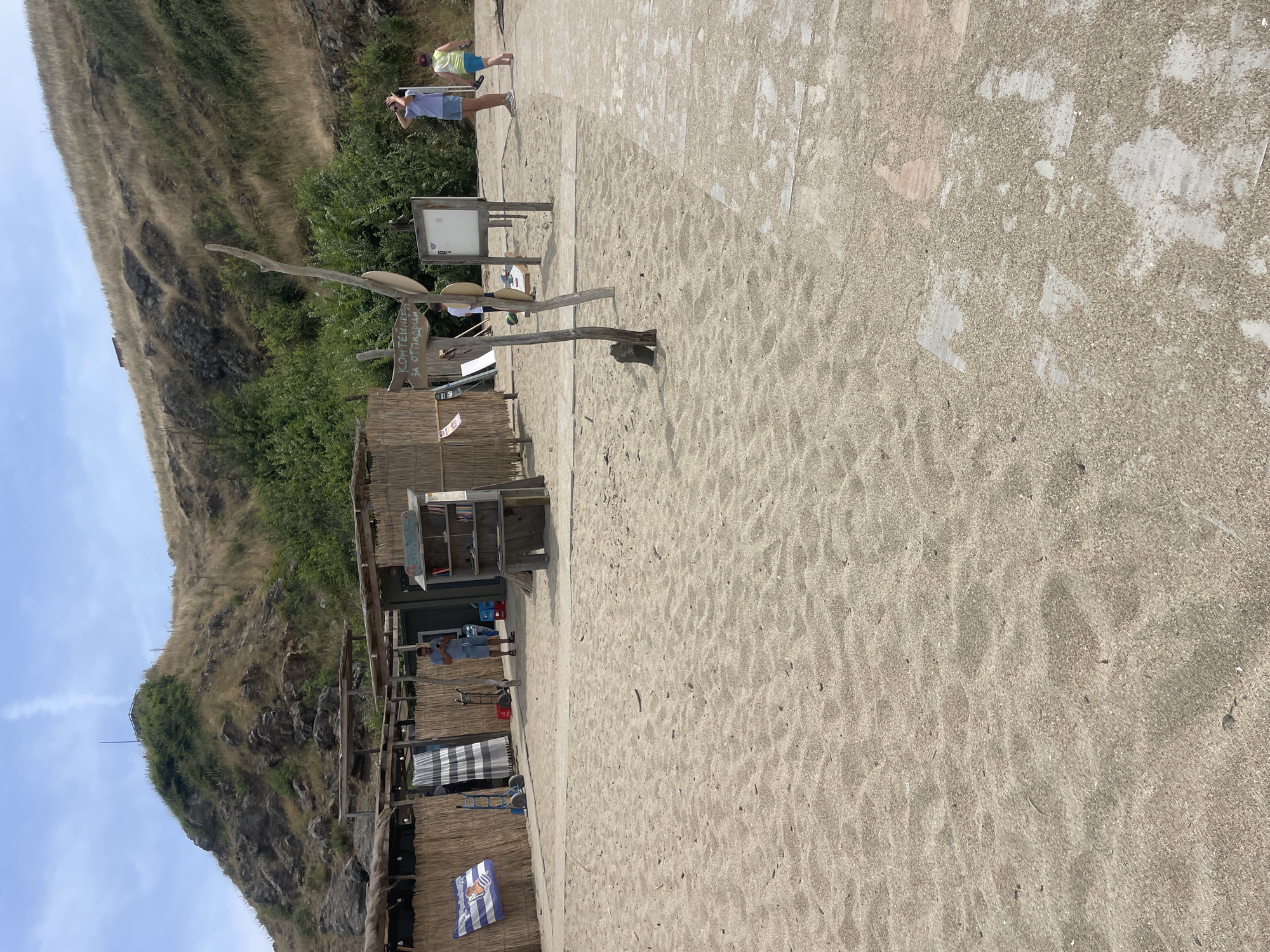 The image depicts a sandy beach scene in Sinemorets, Bulgaria, under a partly cloudy sky. In the foreground, a sandy beach with some scattered debris stretches towards a set of structures made of natural materials like bamboo and straw. These structures appear to be beach amenities, possibly changing rooms or small service areas, with a blue and white striped curtain visible.
In the middle ground, a man in a blue t-shirt and shorts is standing near a wooden bookshelf filled with books. He is looking at his phone. To his right, a signpost with Cyrillic writing, possibly indicating "containers for waste," is planted in the sand. Further to the right, two children are present. One child, wearing a blue t-shirt and shorts, is looking up at the sky, possibly taking a photo with their phone. Another child in a green t-shirt and blue shorts is walking away from the camera. A wooden board with some information or art is mounted on a post near the signpost.
In the background, a steep, grassy hill with rocky outcrops rises above the beach structures. The hill is covered in dry grass and some scattered bushes. The sea is not visible in this frame. The overall impression is a relaxed, natural beach environment with basic amenities and a few visitors enjoying the day.