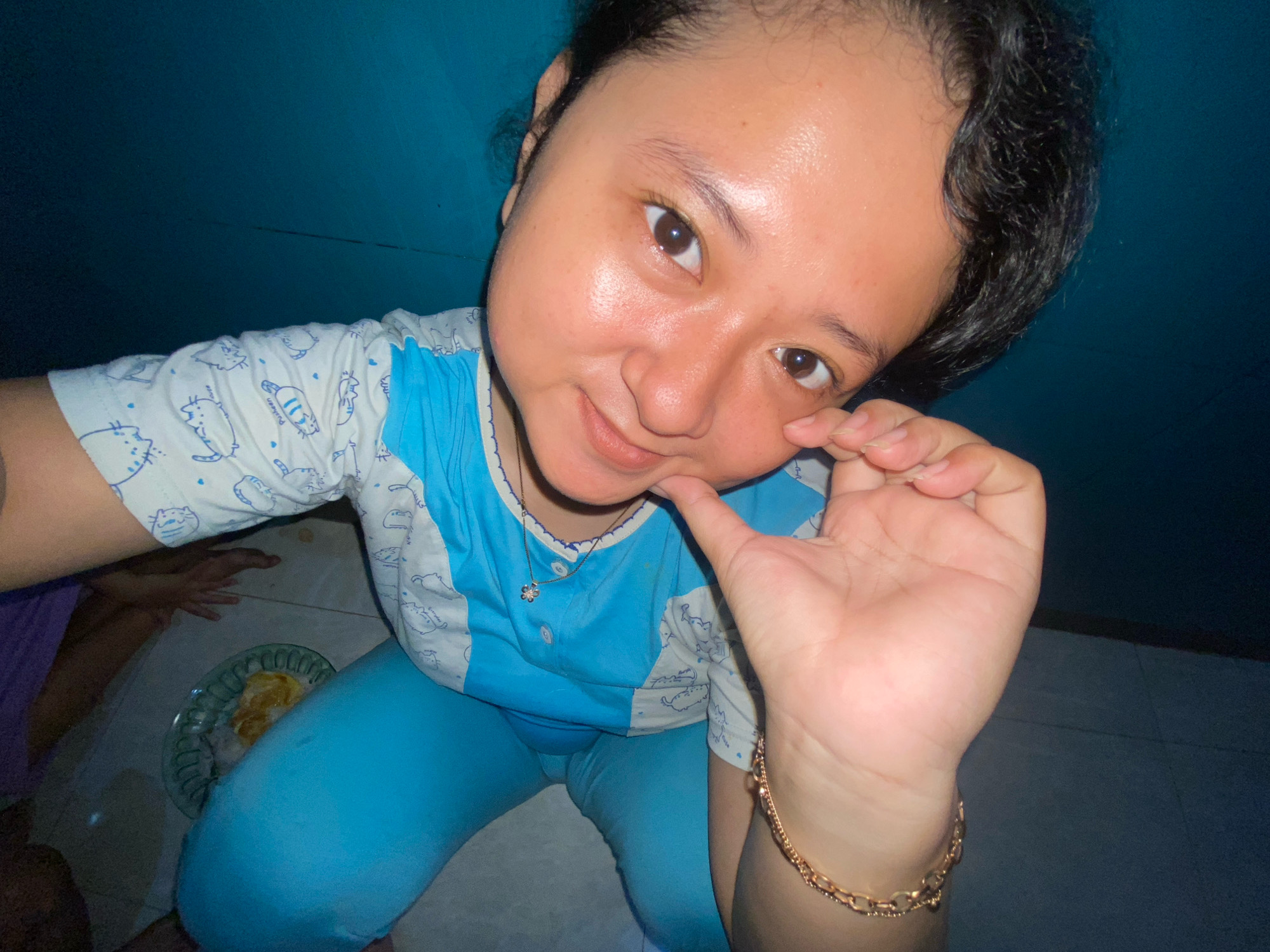 A young female is taking a selfie in what appears to be a dimly lit room, possibly a bedroom, with a blue wall in the background. She is wearing a light blue t-shirt with a white pattern of cats and blue pajama pants. The t-shirt has a small silver flower pendant necklace. She is resting her chin on her right hand, and her left hand is raised, with her fingers gently touching her face. A gold bracelet is visible on her left wrist. In the lower left corner of the image, a green plate with some food, possibly rice and eggs, is visible on the floor. Another person, partially visible in a purple garment, is in the background. The floor appears to be tiled. The scene is consistent with a personal space within a dwelling in the city of Gelam, Indonesia.