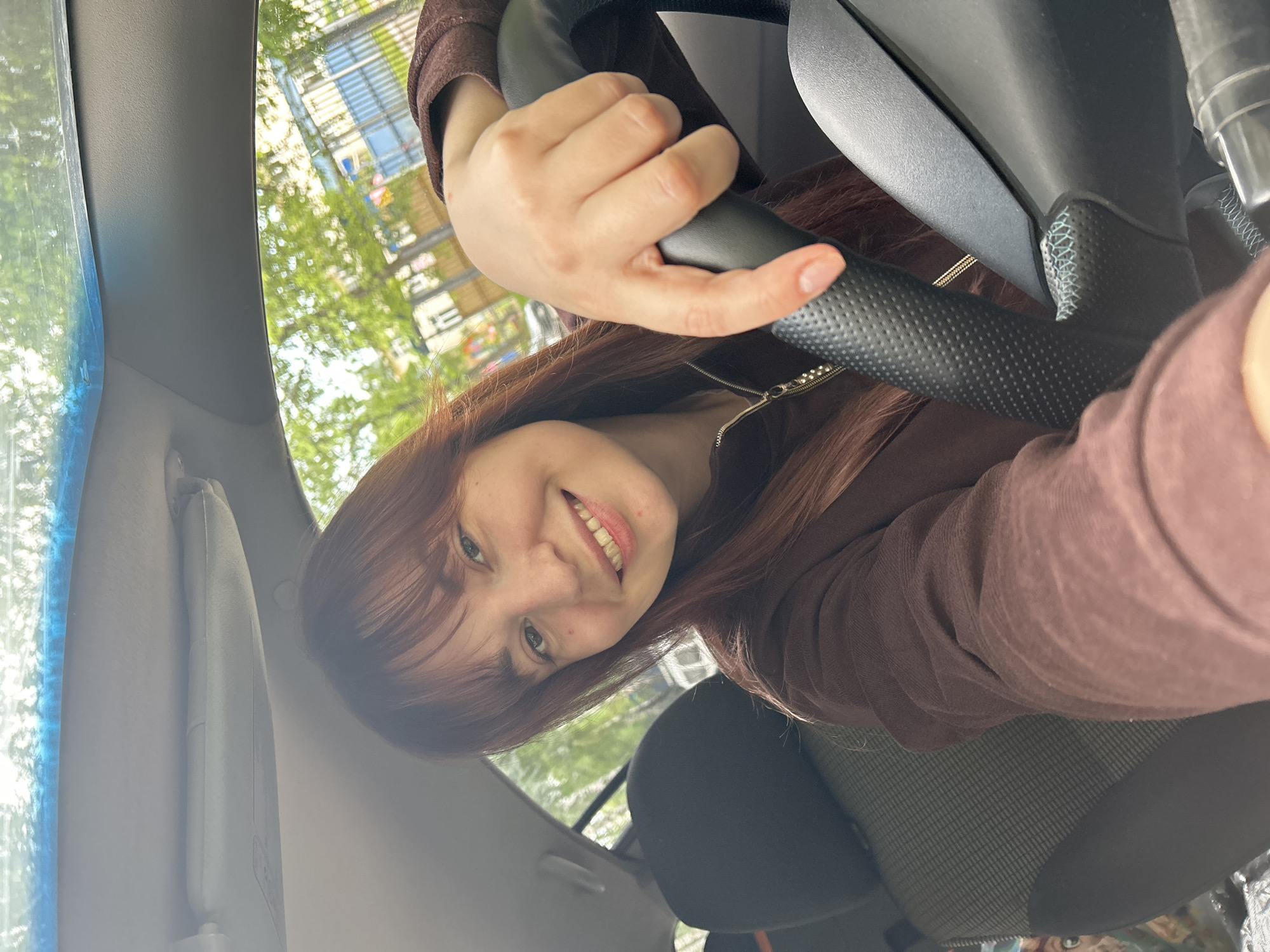 A selfie taken from inside a car shows a young woman smiling at the camera. She has long reddish-brown hair with bangs and is wearing a brown long-sleeved top, possibly a zip-up hoodie or jacket. Her left hand is resting on a black, textured steering wheel, suggesting she is in the driver's seat.
The car's interior features a dark headrest, a patterned car seat fabric below it, and a grey sun visor. Through the car windows, a bright outdoor scene is visible. Green trees with lush foliage and a building with horizontal elements, possibly balconies or blinds, can be seen in the background, indicating an urban or suburban setting. The reflections on the windshield show bright light, suggesting it is daytime and likely sunny. The woman's expression is joyful, characterized by a wide smile revealing her teeth. No discernible text is visible within the image. The location context provided is Chaikovsky, Russia.