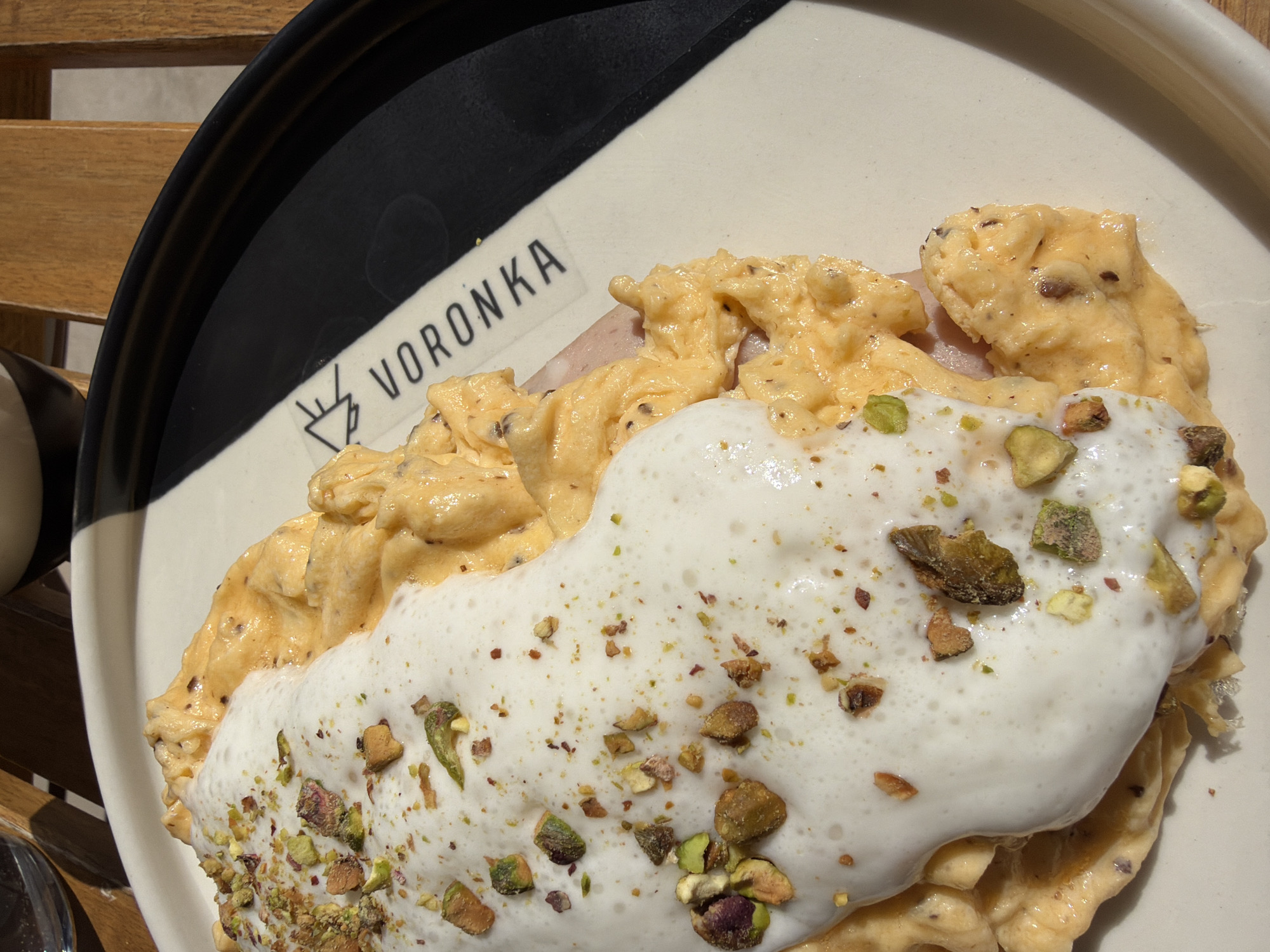 A close-up shot captures a gourmet dish served on a distinctively designed plate, resting on a slatted wooden table. The main subject is a creamy, golden-yellow food item, resembling scrambled eggs, layered beneath a generous topping of frothy white foam. This foamy layer is adorned with an array of chopped green pistachios and smaller, reddish-brown specks, possibly spices or additional nuts.
The plate itself is round, predominantly white, with a striking black band that runs along a portion of its rim. A visible section of the white rim features the brand name "VORONKA" printed in bold black capital letters. Adjacent to the text is a small, stylized graphic, appearing to be a minimalist illustration of a coffee cup or a drip filter symbol.
The setting appears to be outdoors or in a brightly lit space, indicated by the sharp shadows cast by the plate and the food onto the wooden table, suggesting strong daylight, likely morning or early afternoon. A sliver of a dark cup or container is visible on the far left edge of the frame. This culinary presentation, featuring carefully prepared ingredients, hints at a pleasant dining experience in Boreti, Montenegro.