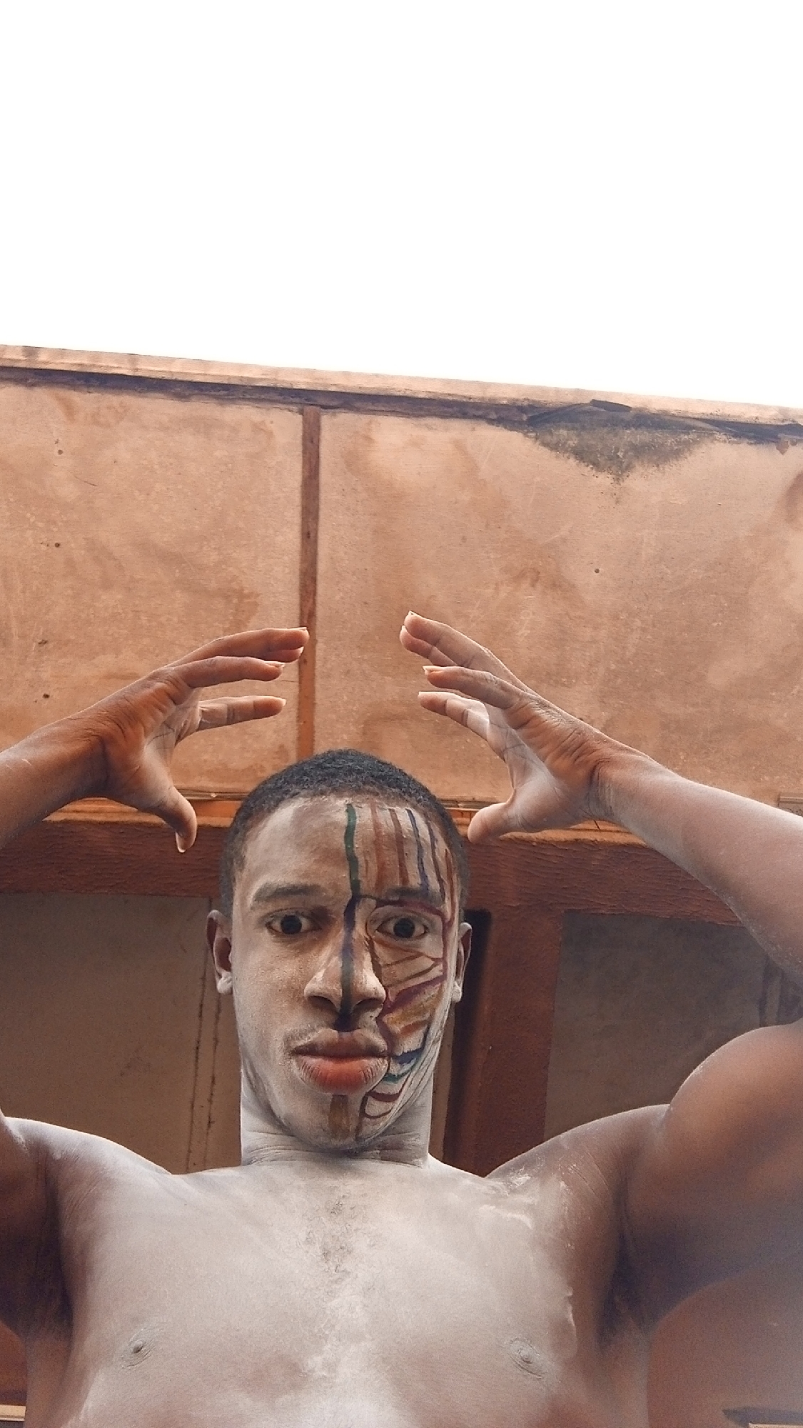 A person with dark skin is viewed from a low angle. The person has their upper body exposed and has white powder on their chest and neck. Their face is painted with two different designs, one side is primarily brown and the other is a mix of greens, reds, and browns. The person has both hands raised with palms facing inwards, and their fingers are slightly curved. Behind the person is a wall of wooden or composite boards. Above the wall is a pale sky. The location is Unknown, Unknown.