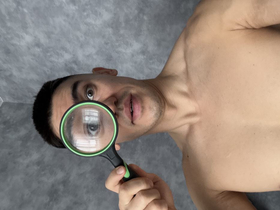 A bare-chested man holds a green-rimmed magnifying glass to his eye. The glass reflects what seems like a window. He gazes directly at the viewer with an intrigued expression. He is positioned from a low angle, emphasizing his chest and face. The background is a grey, mottled surface.