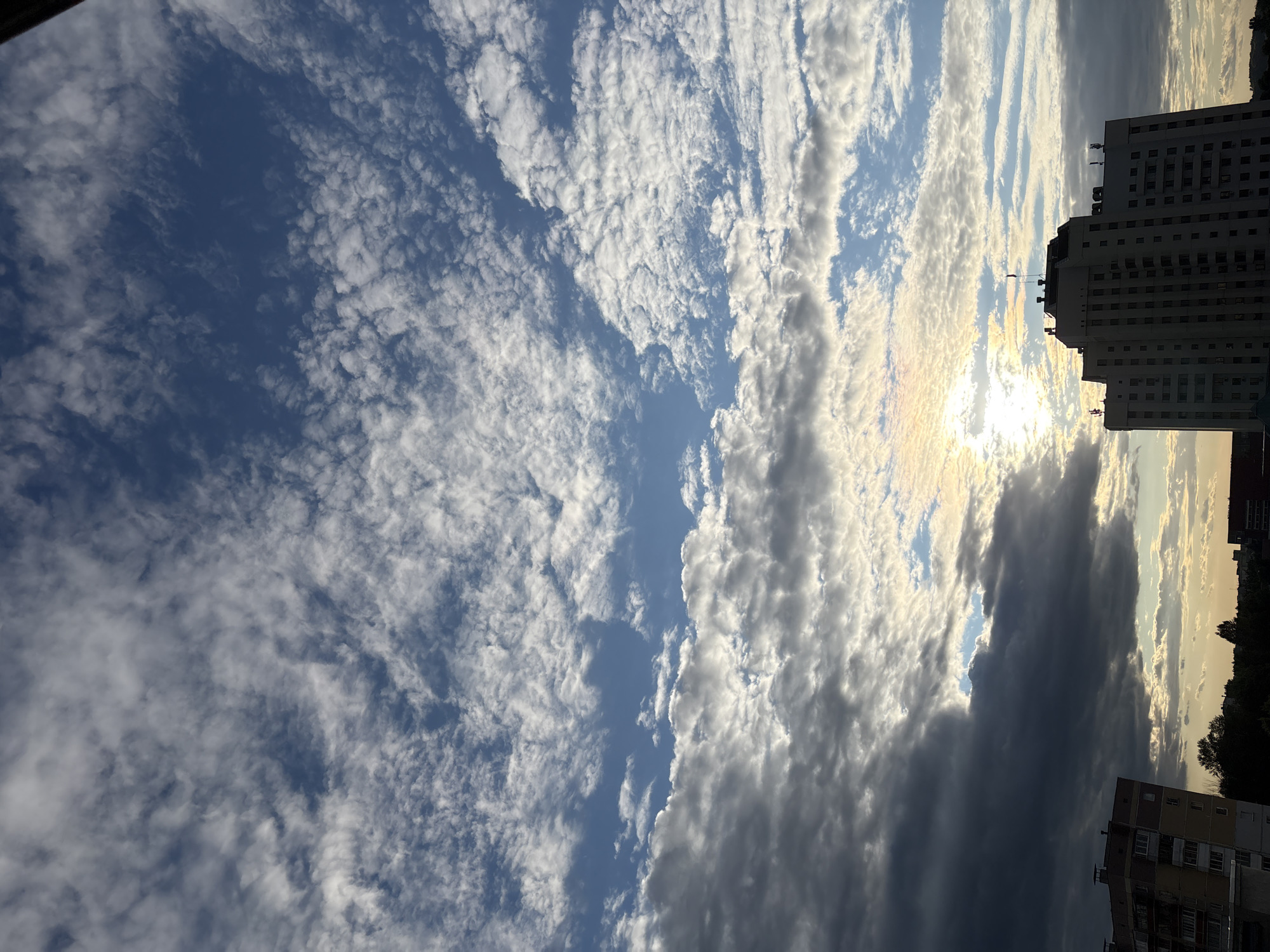 The image depicts a sky filled with dramatic clouds, with a large, multi-story building silhouetted against the bright sky in the foreground. The building appears to be a high-rise apartment complex or office building, with numerous windows visible. A smaller building is visible on the left side of the frame. The sky features a mix of dark, heavy clouds and lighter, wispy clouds, with the sun shining through in the center of the image, creating a halo effect. The overall impression is of a dynamic sky, possibly during sunset or sunrise. Based on the architectural style of the buildings, which are typical of Soviet-era construction, and the presence of a flag on top of the larger building, this scene is likely located in a city in Belarus, potentially Vitsebsk, given the prompt. There are no people visible in the image.