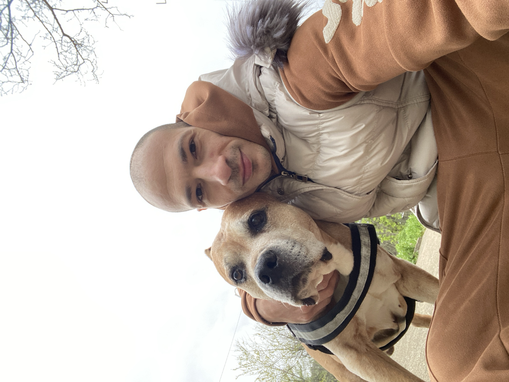 A selfie captures a man and a dog. The man has a shaved head, light skin, and is smiling at the camera. He is wearing a light brown hooded sweatshirt and a light colored puffy vest with a fur-lined hood. The dog, held by the man, has light tan fur and is wearing a black and gray harness. The background is blurred, showing hints of trees and sky. While location is not definitively identifiable, the photo was provided with the assumption it's in Pleven, Bulgaria. If this is correct, the setting appears to be an outdoor public area in Pleven. The interaction suggests a bond between the man and his dog.