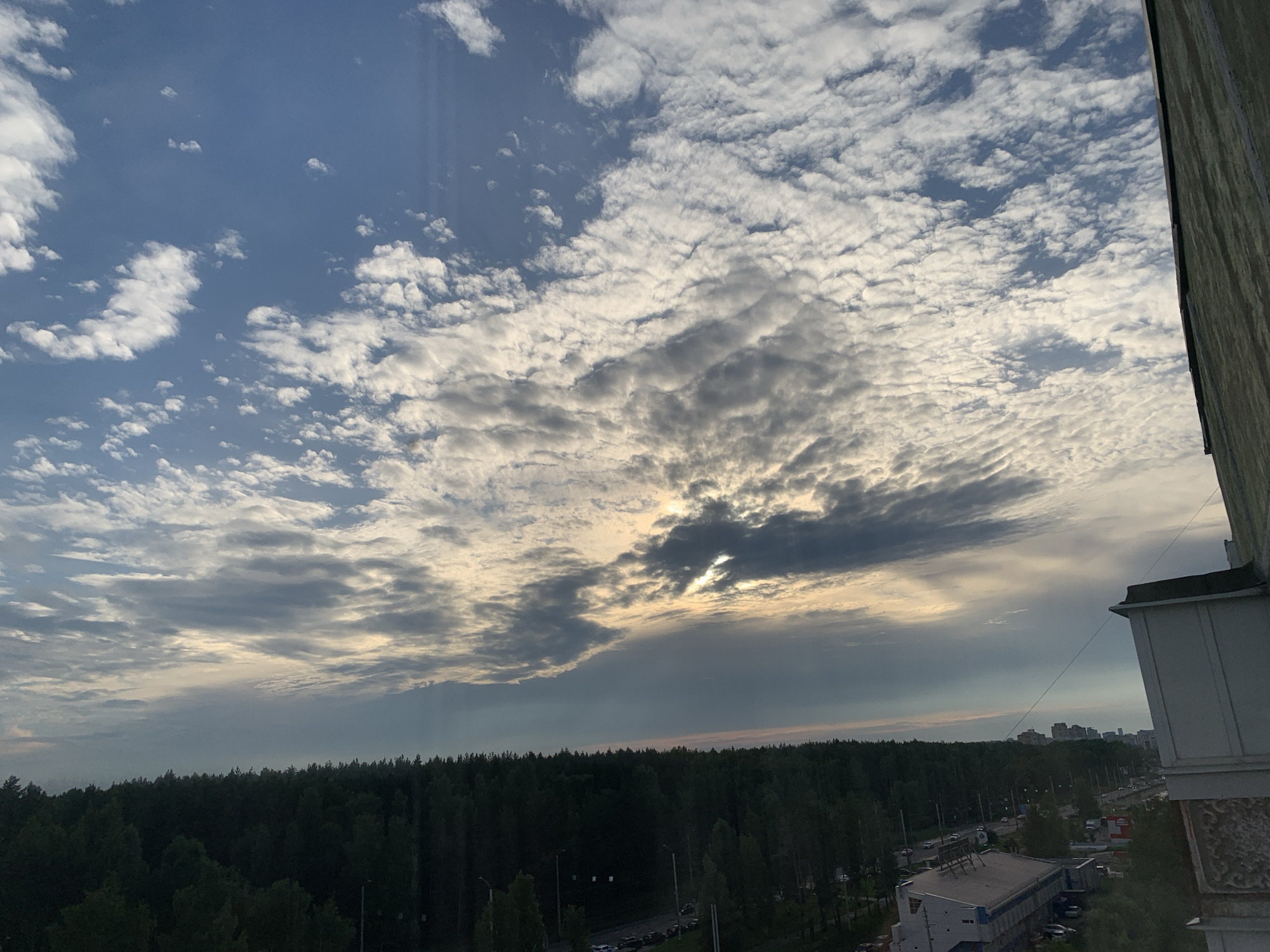 This image, taken from a high vantage point in the city of Ivanovo, Russia, depicts a panoramic view of the sky, a dense forest, and a portion of the urban landscape. The sky is a dynamic canvas of clouds, with patches of clear blue interspersed with numerous white, puffy cumulus clouds and darker, more textured altocumulus formations. The sun is partially obscured by a band of clouds, creating a soft, diffused light that illuminates the scene with a warm, golden glow.
Below the sky, a vast expanse of dark green forest stretches across the horizon, its trees forming a uniform, dense canopy. This natural boundary separates the sky from the urban elements. To the right of the forest, a road with several cars and streetlights is visible, suggesting a moderately busy street. In the distance, several multi-story buildings can be seen, indicating the presence of a residential or commercial area within the city. A section of a building's facade, likely from the vantage point of the photographer, is visible in the foreground on the right side of the image, characterized by concrete and architectural details. No people are discernible in the image. The overall impression is one of a transitional zone between nature and urban development under a dramatic sky.