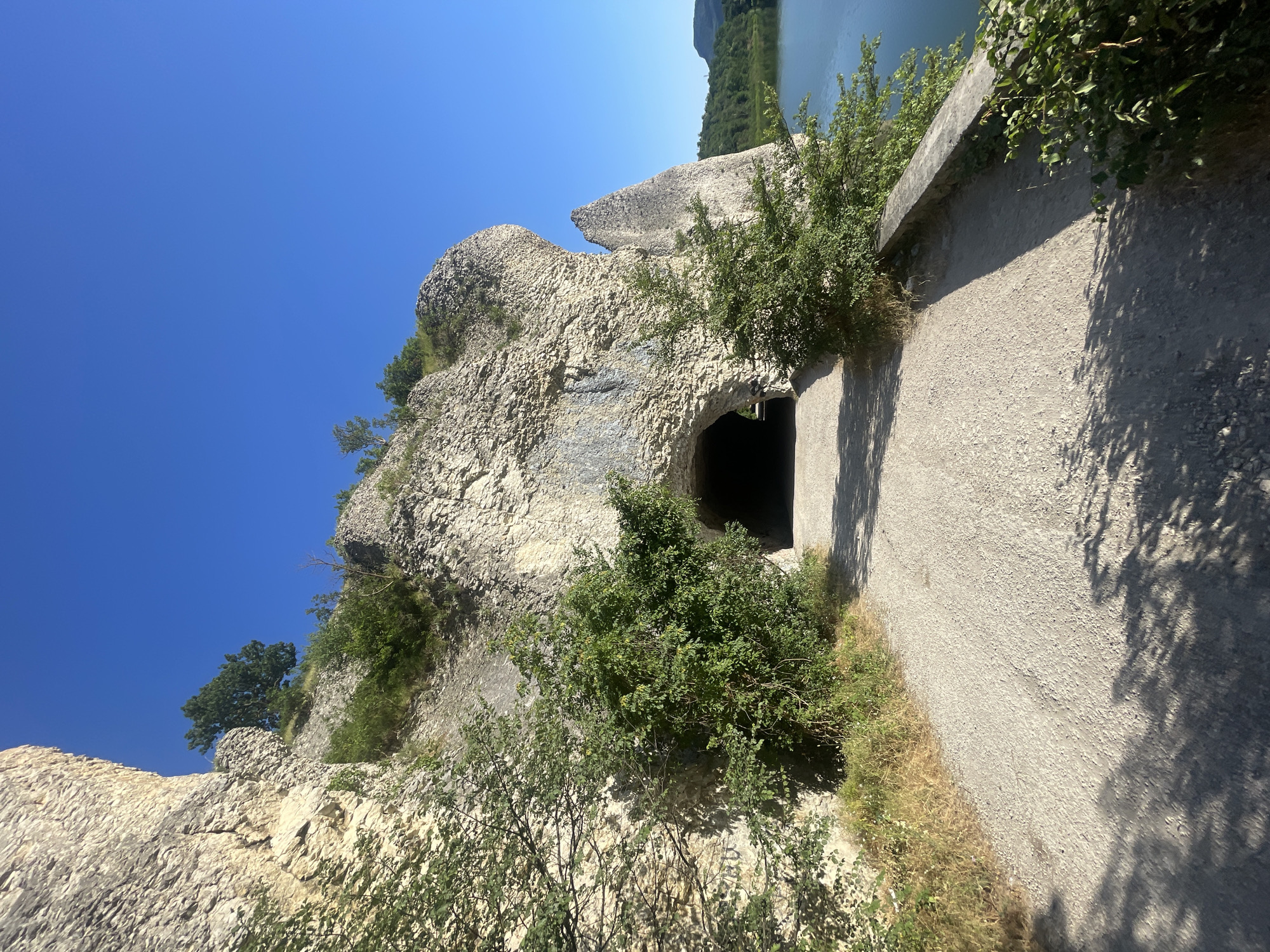 The image depicts a sunny day at a rocky cliff face with an opening that appears to be a tunnel or cave entrance. A paved path runs alongside the cliff. Green bushes and trees are growing on and around the rocks, and the path is lined with dry grass and shadows cast by foliage. In the distance, a body of water, likely a lake or river, is visible, bordered by lush green trees. The sky is a clear, vibrant blue. The scene suggests a natural, outdoor environment, possibly a hiking trail or a scenic viewpoint. There are no people or specific objects that indicate a particular activity, but the setting evokes a sense of exploration or peaceful contemplation. The time of day is clearly midday due to the strong sunlight and the sharp shadows. The location cues of the rocky terrain, water, and vegetation are consistent with a natural landscape, and the overall impression is one of a serene summer day. No text is visible in the image.