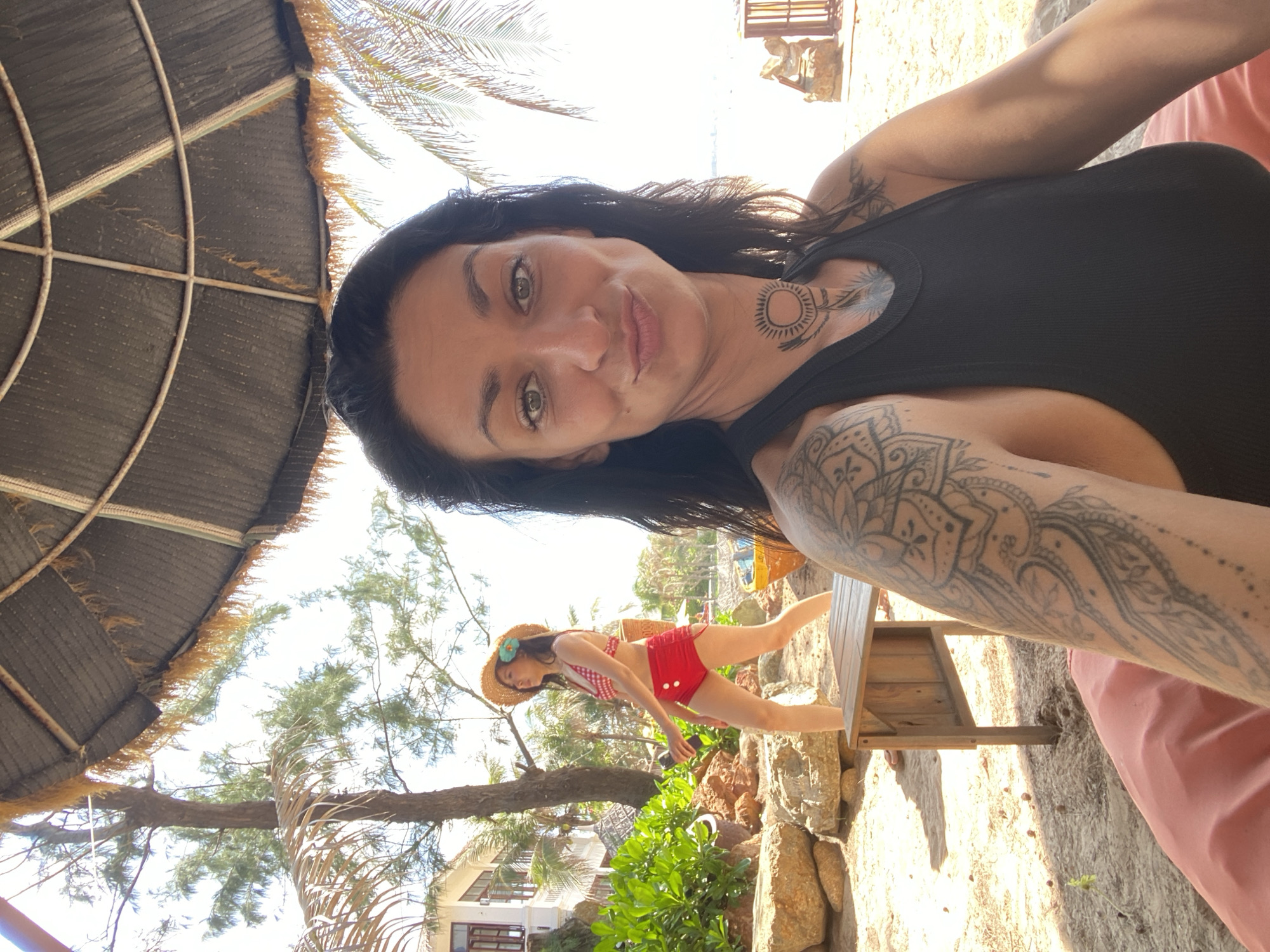 The image depicts a selfie taken from a low angle, showing a woman with long dark hair and a black tank top in the foreground. She has visible tattoos on her arm and neck. Behind her, another woman wearing a straw hat, a red and white checkered bikini top, and red high-waisted shorts is walking to the left, holding a dark object. The background reveals a beach scene with palm trees, lush green foliage, rocks, a wooden table, and a thatched umbrella. The location is identified as Phu Thuy Ward, Vietnam, suggesting a tropical or subtropical environment. The overall setting appears to be a beach resort or recreational area.