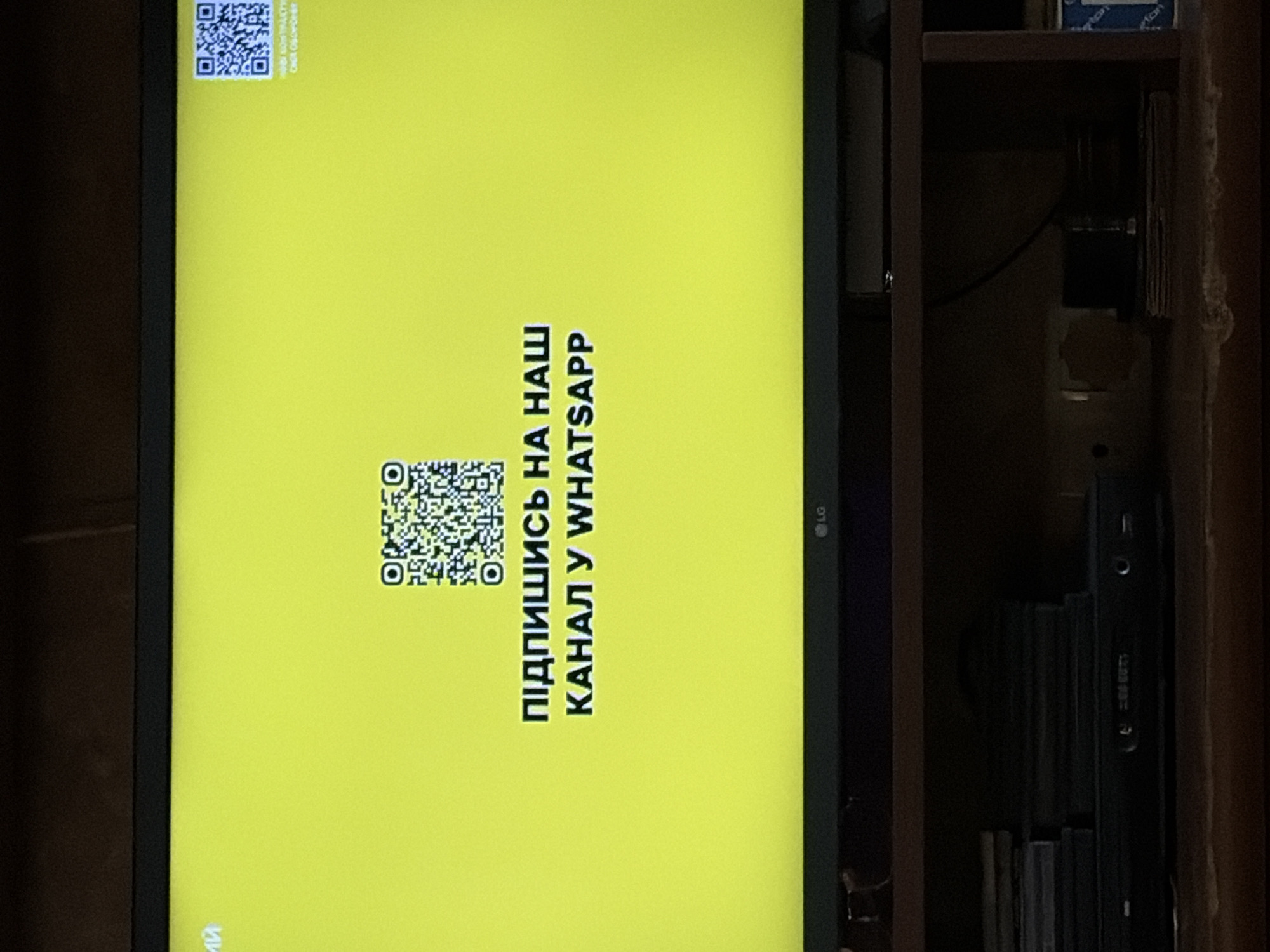 The image is a close-up, slightly angled shot of a television screen displaying a bright yellow background. In the center of the screen, there are two QR codes. The left QR code is larger and is positioned slightly to the left of the center. The right QR code is smaller and is located to the right of the center, closer to the edge of the screen. Below the QR codes, in bold, white, uppercase letters, is the text "ПІДПИШИСЬ НА НАШ КАНАЛ У WHATSAPP". The screen also displays the LG logo in the bottom center. The television is placed on a dark shelf, and below the shelf, a black electronic device, possibly a DVD player or set-top box, is visible. There are also some stacked items, likely discs or cases, on the shelf. The background of the image, outside the television screen, is dark and appears to be a wall. The lighting is dim, with the bright screen being the primary light source. The scene is indoors, with no indication of a specific city or location beyond what is implied by the electronic equipment and indoor setting.