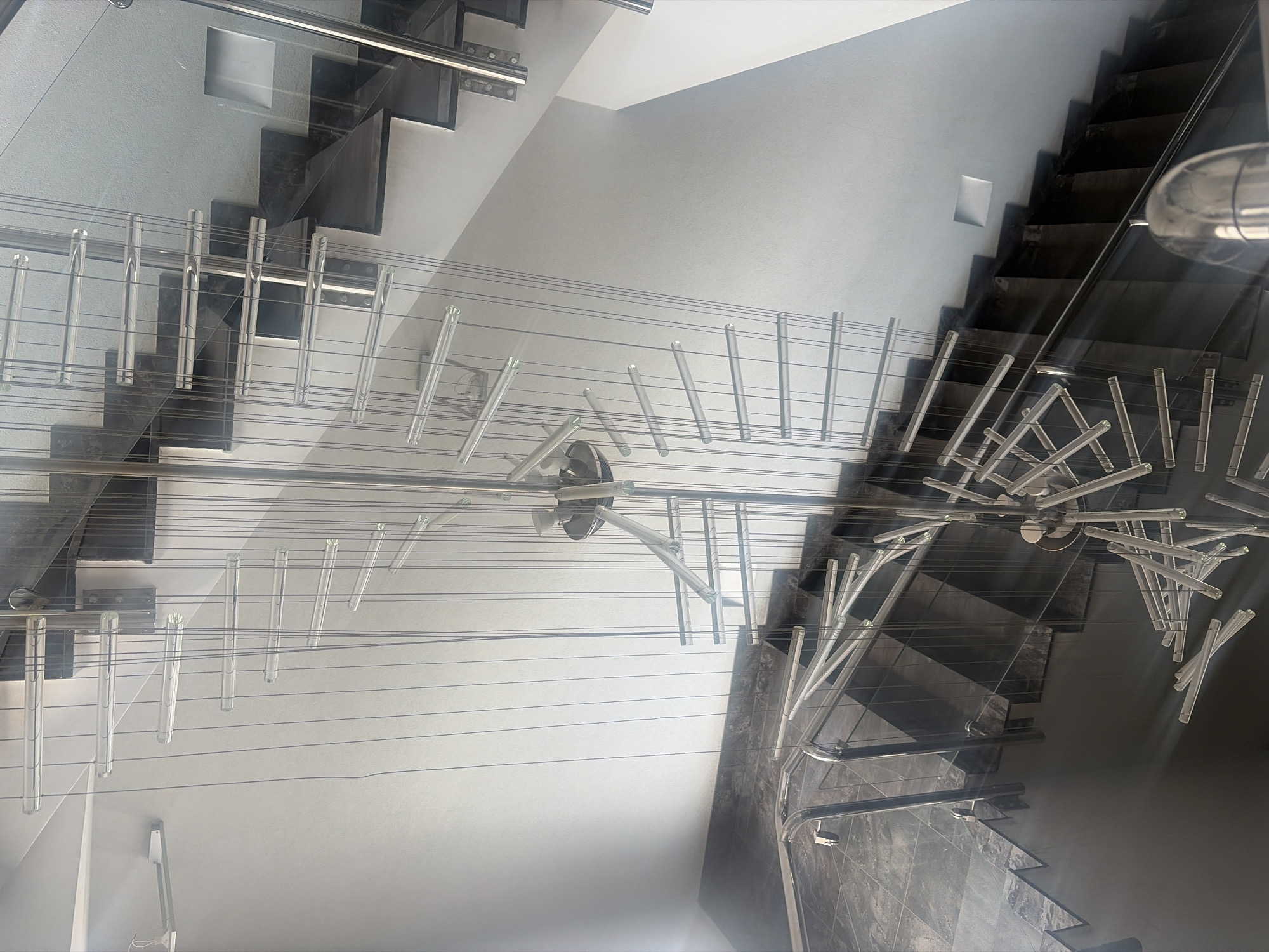 The image shows a view looking down through glass, revealing a staircase with metal railings and a decorative light fixture suspended above. The staircase appears to be in an interior location. The walls are light-colored and the steps are a darker hue. The metal railings feature a sleek, modern design. The decorative light fixture consists of multiple transparent rods suspended from a central point.
The location is in Saint Petersburg, Russia.