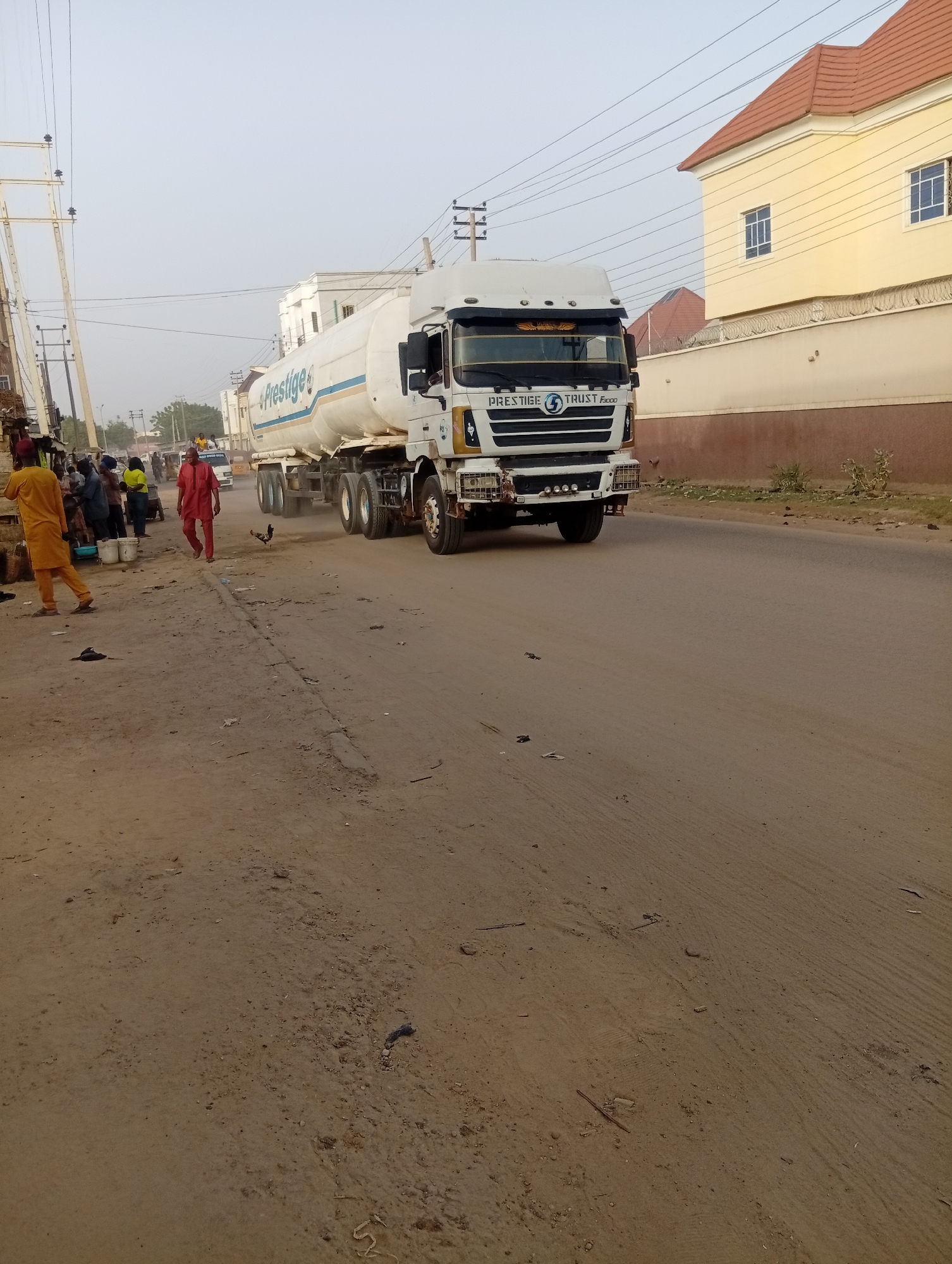 The scene depicts a road in Aviation Quarters, Nigeria. A large white tanker truck, labeled "Prestige Trust," occupies the road, moving from left to right. A group of people is gathered on the left side of the road near some tables and buckets. One man in a red outfit walks along the shoulder. A chicken runs across the road near him. The road is dirt and asphalt, with some debris scattered on the shoulder. A yellow, two-story building with a reddish-brown roof is on the right side of the road. Power lines and poles are visible on the left side of the frame.