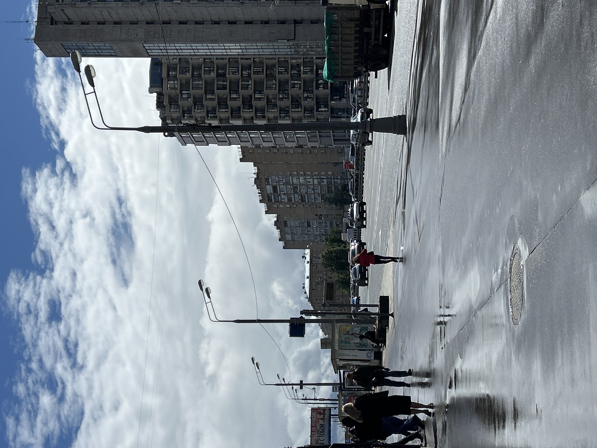The image captures a wet urban street in daylight. The road surface is glossy with reflections, suggesting recent rain. Several people are walking along the sidewalk, some clustered together and others spaced apart. Street lamps line the side of the road, their poles rising into the sky. Buildings of various heights are visible, one of which is a high-rise with numerous balconies. The sky is filled with scattered clouds, with patches of blue showing through. Cars and trucks are parked along the roadside or moving in the distance. The overall impression is of a typical urban scene after a rainfall.