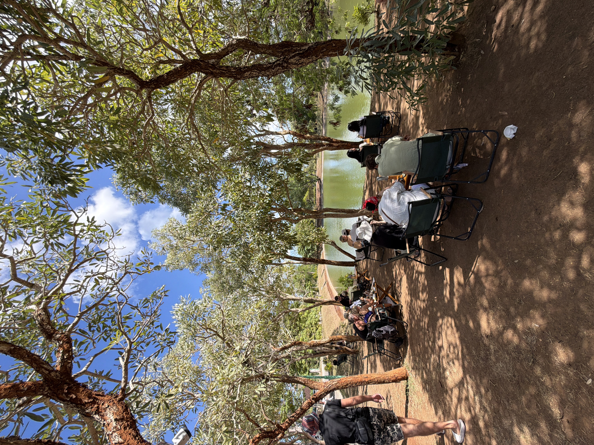 The image depicts an outdoor seating area under trees, adjacent to a calm, green body of water, likely a lake or pond. Several groups of people are seated at tables and in camping chairs, engaged in conversation or enjoying the surroundings. The ground is covered in reddish-brown soil with scattered dry grass and leaf litter. The trees have distinctive reddish-brown bark and sparse green foliage, with sunlight filtering through the branches creating dappled shadows on the ground. In the background, more trees and foliage are visible, with a clear blue sky above dotted with white clouds. One person in the foreground is walking away from the camera, wearing a black t-shirt, patterned shorts, and sandals. The overall scene suggests a relaxed outdoor dining or gathering setting. The location is identified as Kuta Selatan, Indonesia.