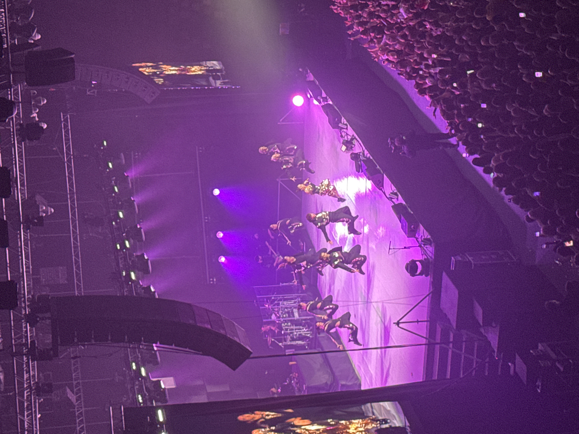 The image depicts a concert stage illuminated by purple lighting, likely located in Perm, Russia. On the stage, a group of performers are visible, clad in coordinated outfits. A large crowd of spectators fills the foreground and background, suggesting a well-attended event. The stage features lighting rigs and sound equipment, common elements in a concert setting.