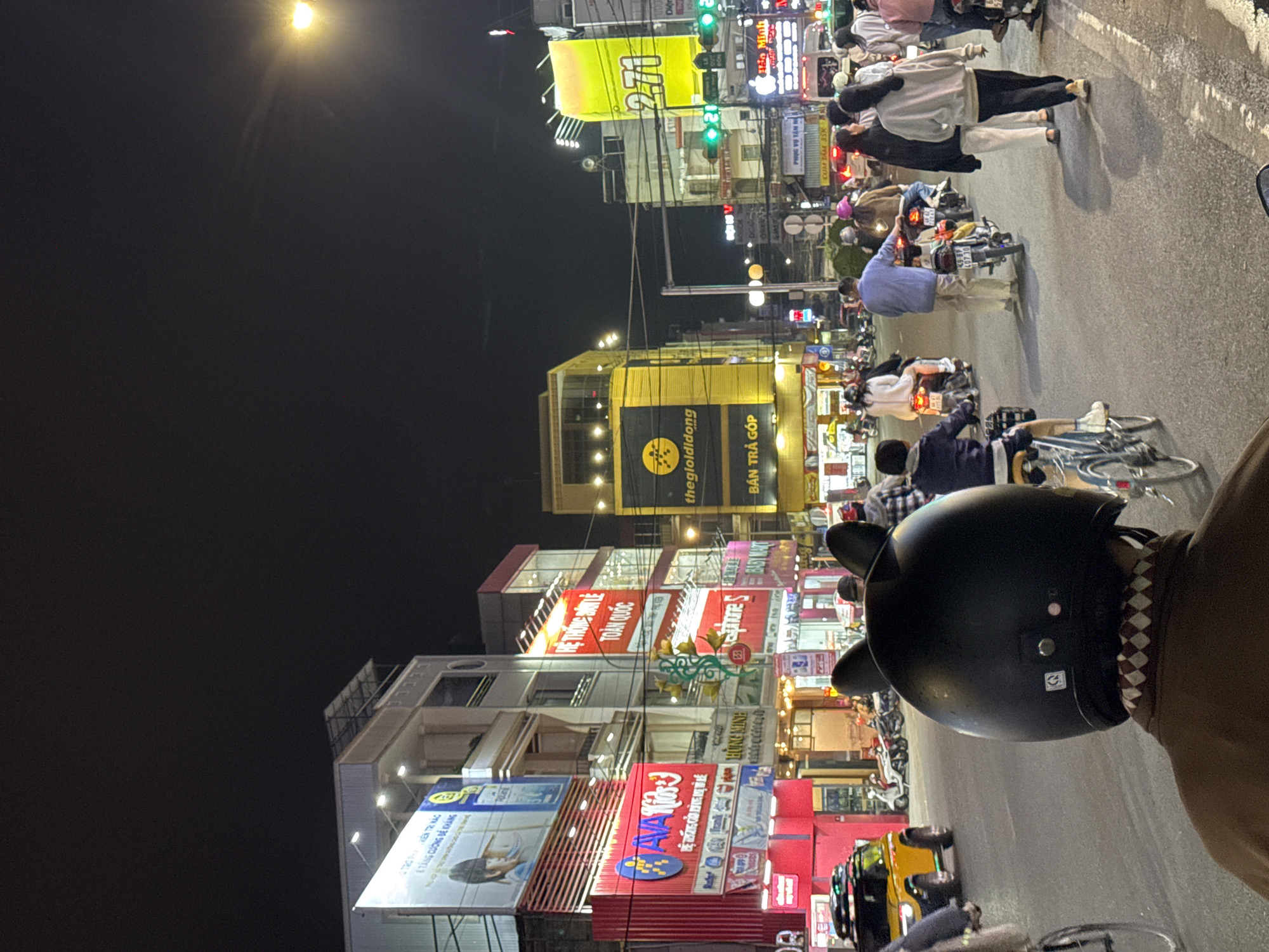 This is a vertical, eye-level shot taken at night in what appears to be a busy street in Da Lat, Vietnam. The scene is dominated by brightly lit buildings adorned with numerous signs in Vietnamese. Many of these signs are advertisements for various products and services, including electronics stores, children's items, and promotions like "BÁN TRẢ GÓP" (installment sales). The street is filled with people, mostly on motorcycles, suggesting a lively evening atmosphere. Some individuals are walking, while others are riding or stopped on their bikes.
The lighting is primarily artificial, emanating from the buildings, streetlights, and the headlights of vehicles. A prominent yellow neon sign with the number "271" is visible on one of the buildings, as are signs for "thegioididong" (likely a mobile phone and electronics retailer) and "VAVA Kids." The presence of a yellow taxi or similar vehicle adds to the urban street scene. The overall impression is one of a vibrant and active commercial district at night. The image seems to be taken from the perspective of someone on a motorcycle, with a black helmet with ear-like decorations visible in the foreground, partially obscuring the view. The weather appears to be clear, as indicated by the sharp and bright artificial lights.