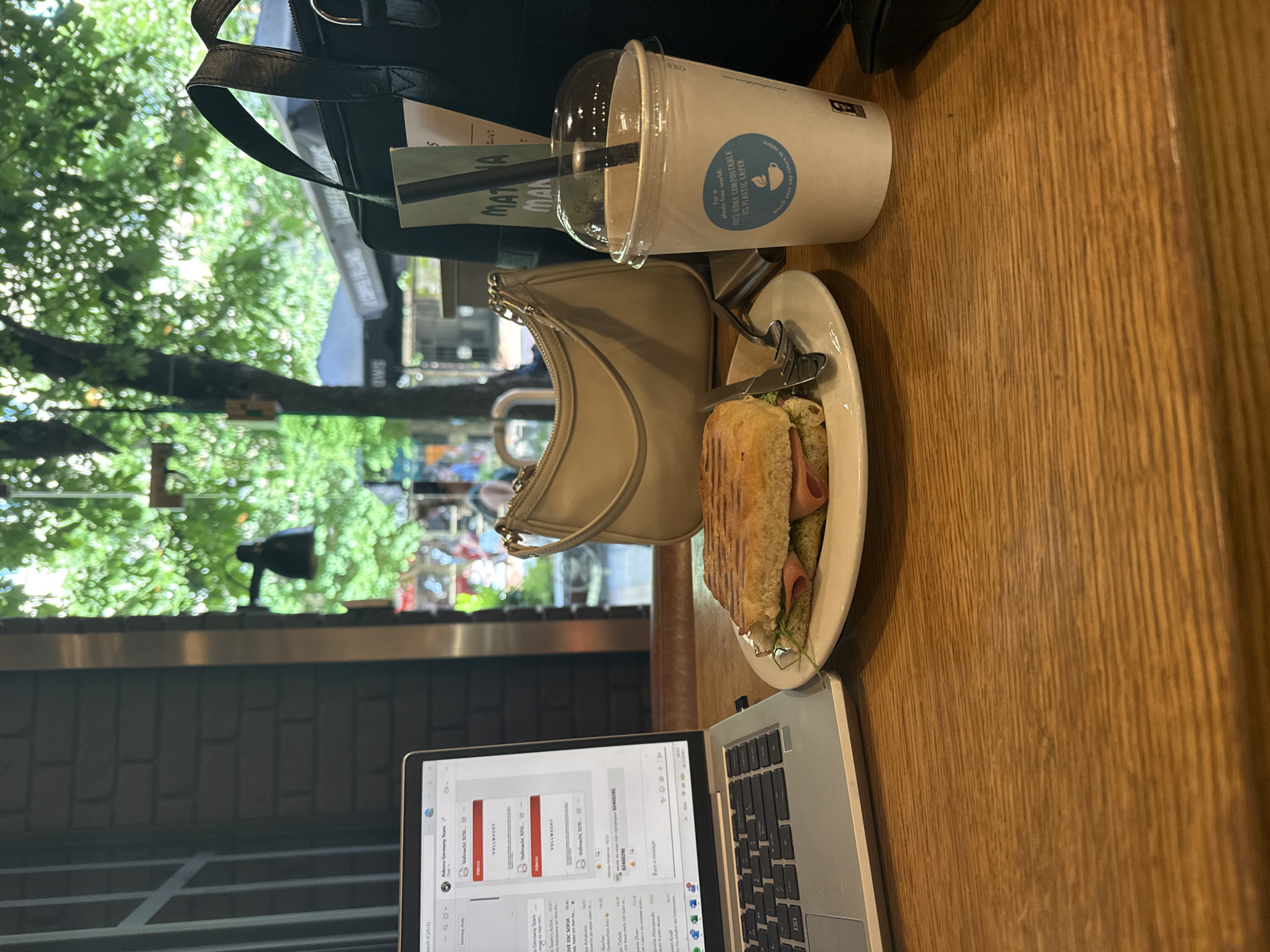 A slightly canted medium shot, taken from above, depicts a casual workspace on a wooden table, likely within a cafe, judging by the partial view outside of trees, buildings, and pedestrians. Dominating the composition on the right side is a partially eaten sandwich on a white plate, accompanied by silverware, and a plastic-lidded beverage cup with a straw. A beige shoulder bag is positioned next to the plate. In the foreground, a laptop displays an open application, seemingly related to work. The visible text in the application includes "Adecco Germany Team," "Vollmacht," and other foreign language phrases. The scene also features a black handbag, a menu, and the partially visible outdoor view of what looks like the city of Sofia, Bulgaria. The light suggests it is daytime, and the location is probably a cafe or restaurant in the downtown area.